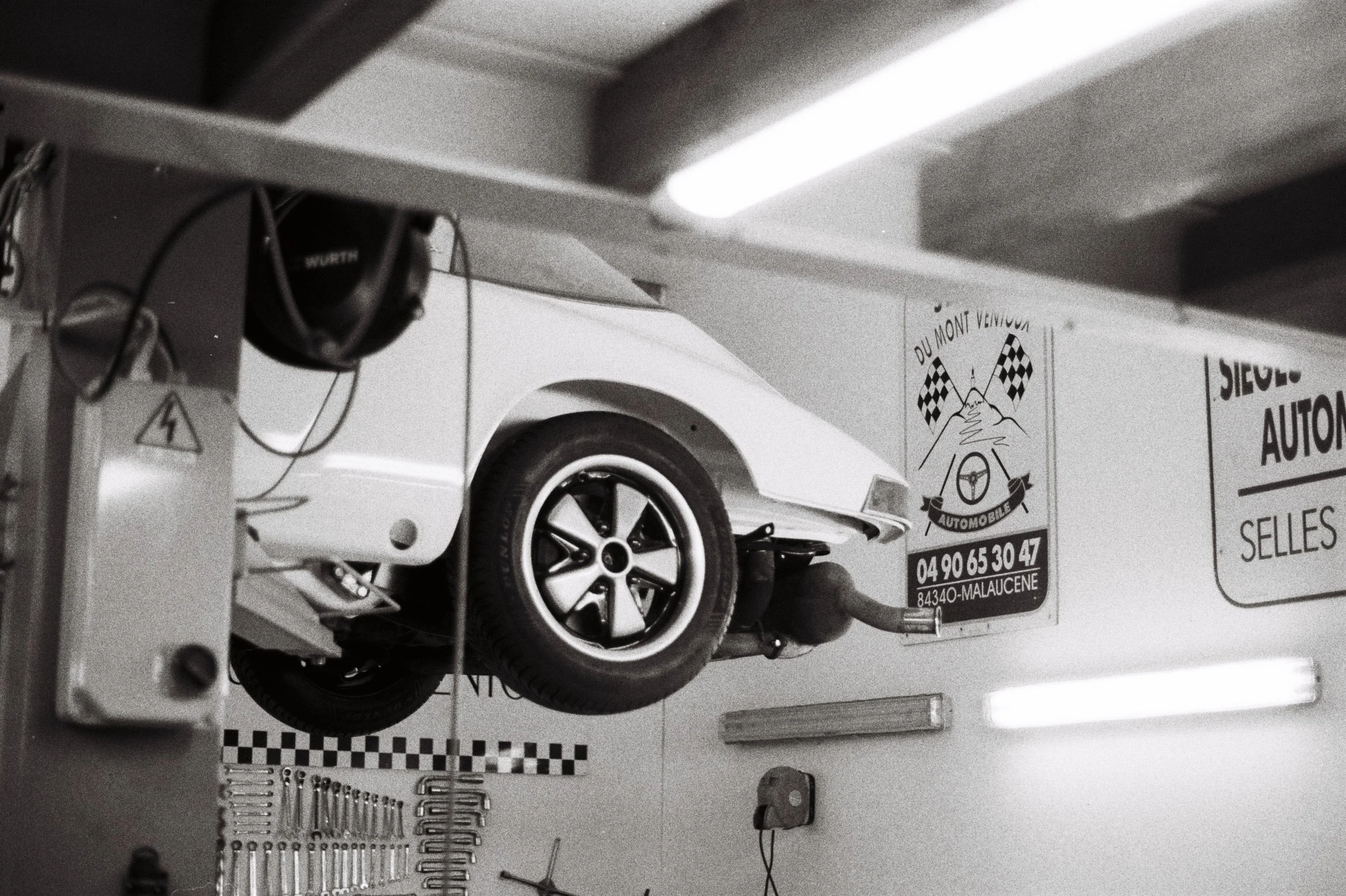 A white vintage sports car mounted on a lift inside an auto repair garage with signs on the wall, black and white checkered banners, and overhead fluorescent lighting.
