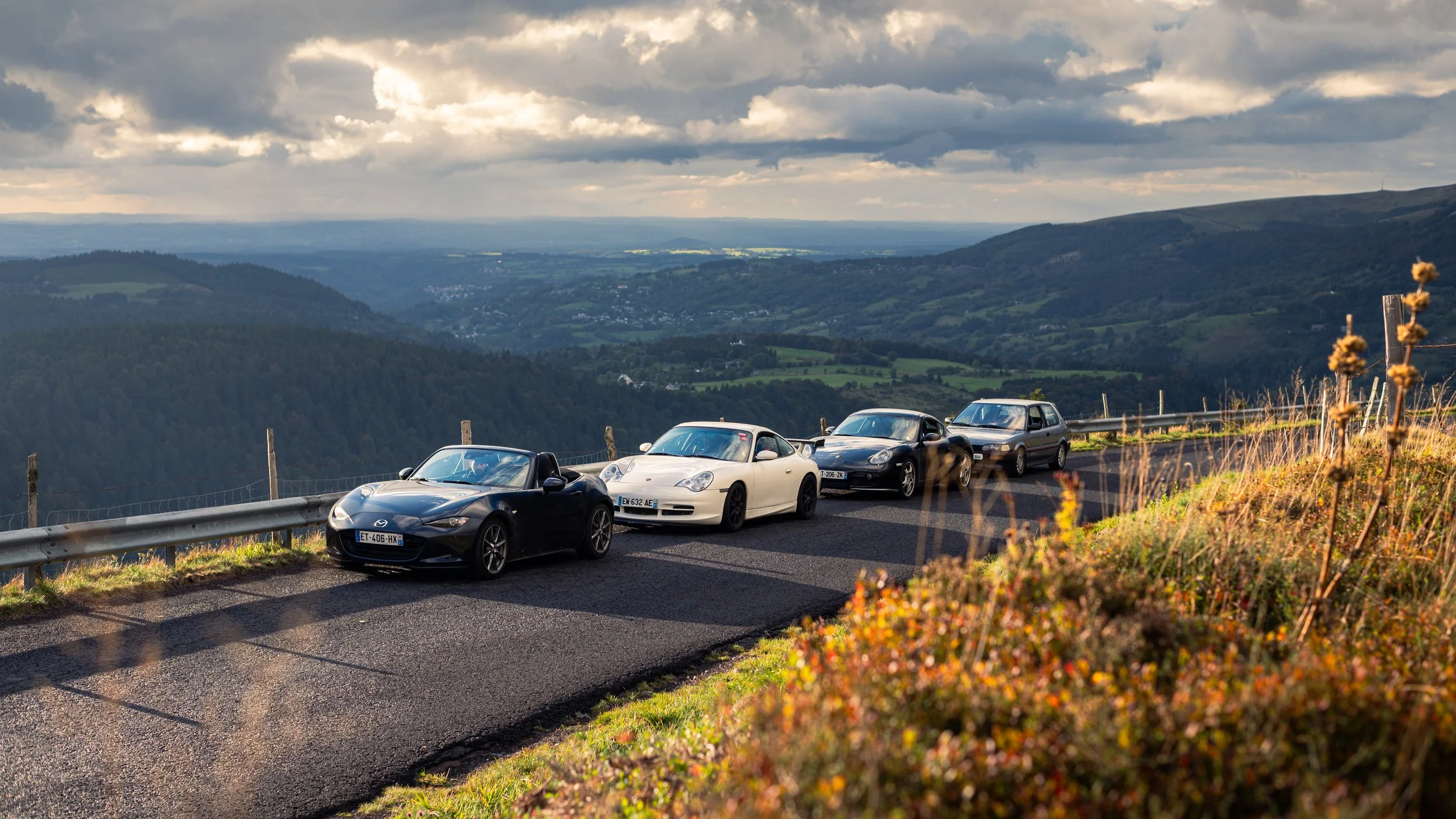Four cars parked along a mountain road overlooking a valley with hills and cloudy sky in the background.