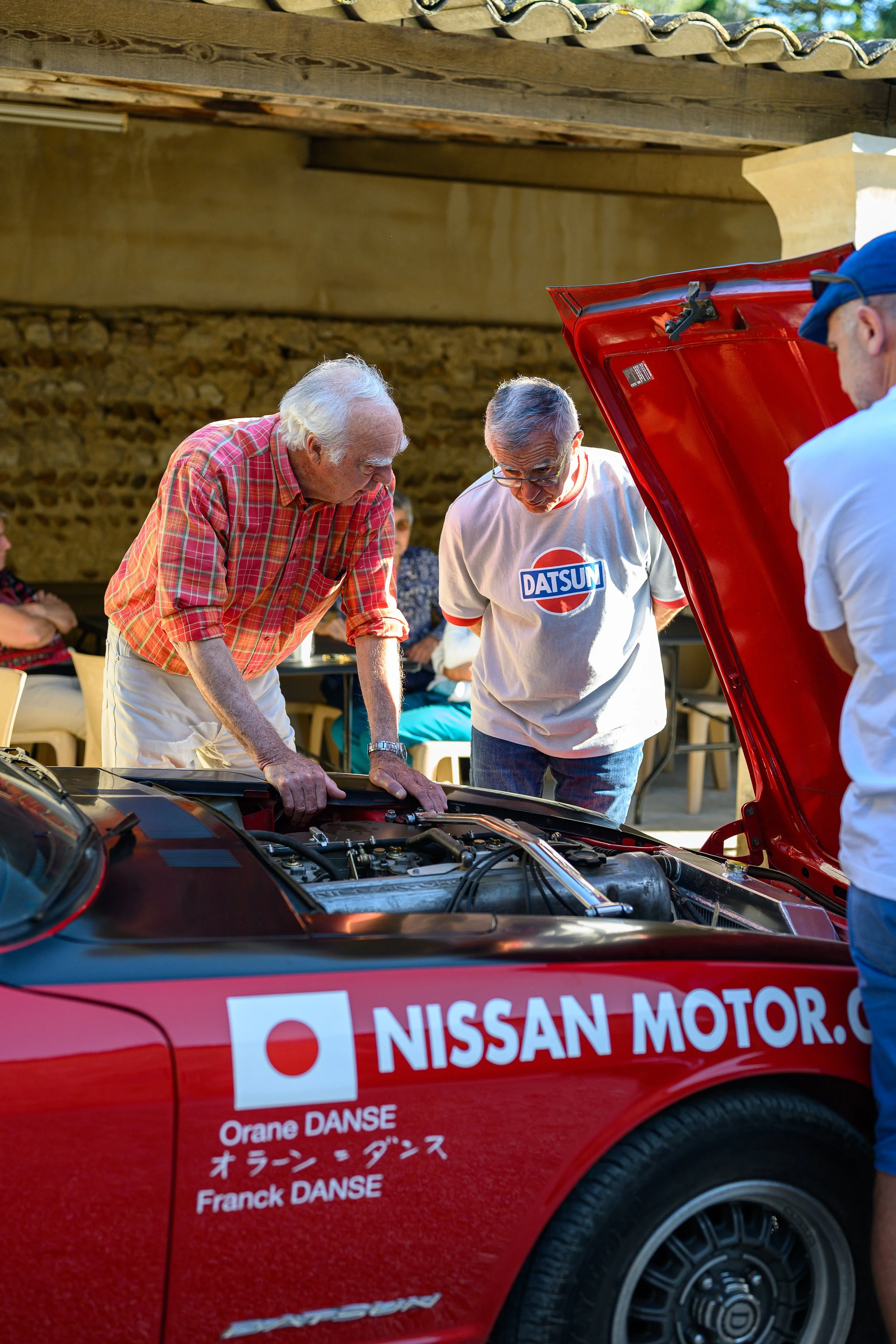 Three men examining the engine of a red Nissan sports car with a hood open at an outdoor event. One man is wearing a plaid shirt, and another is wearing a white T-shirt with a Datsun logo.