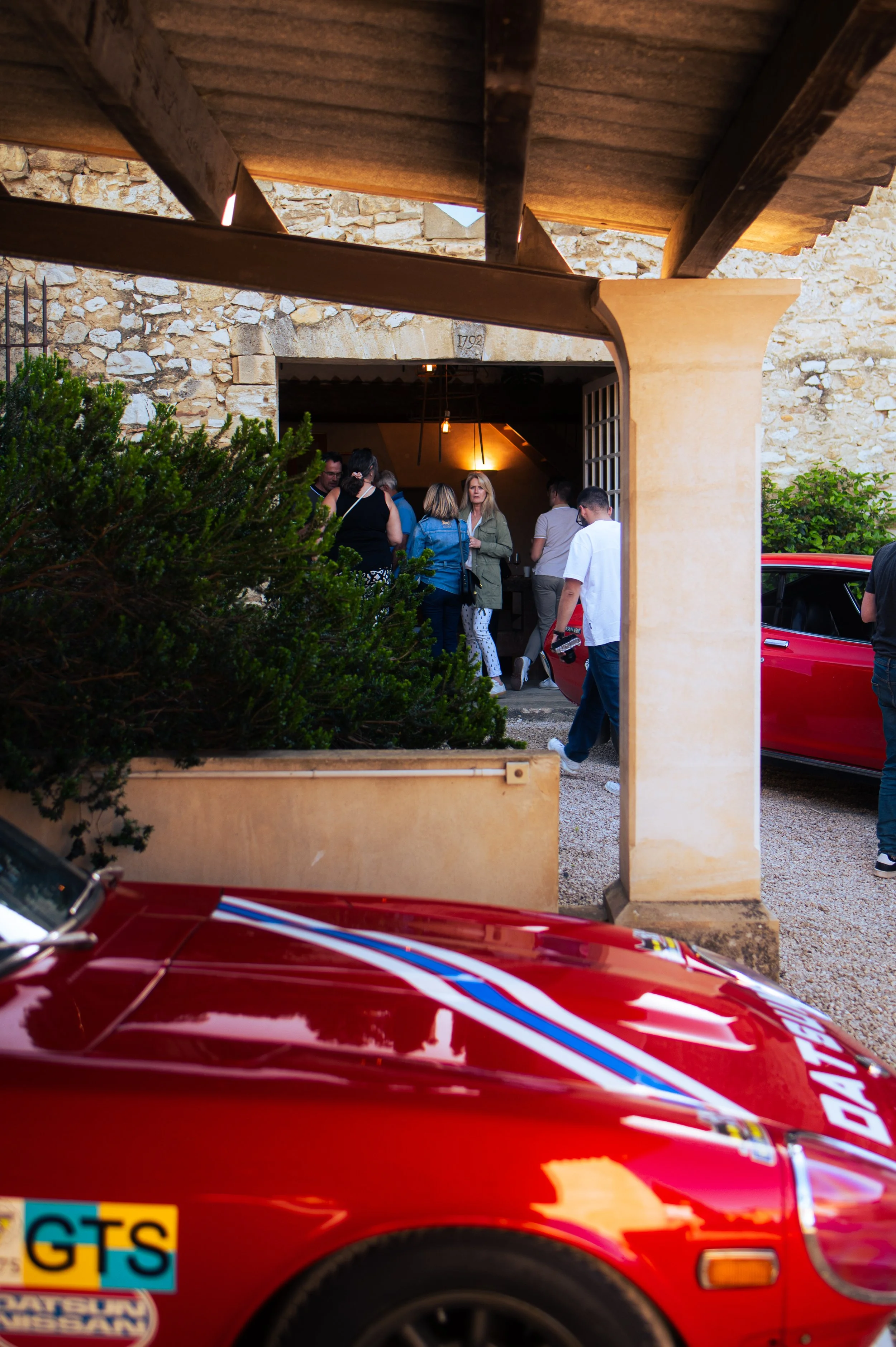 People gathering outside a stone building with a winery door, with a red Datsun 240Z partially visible in the foreground.
