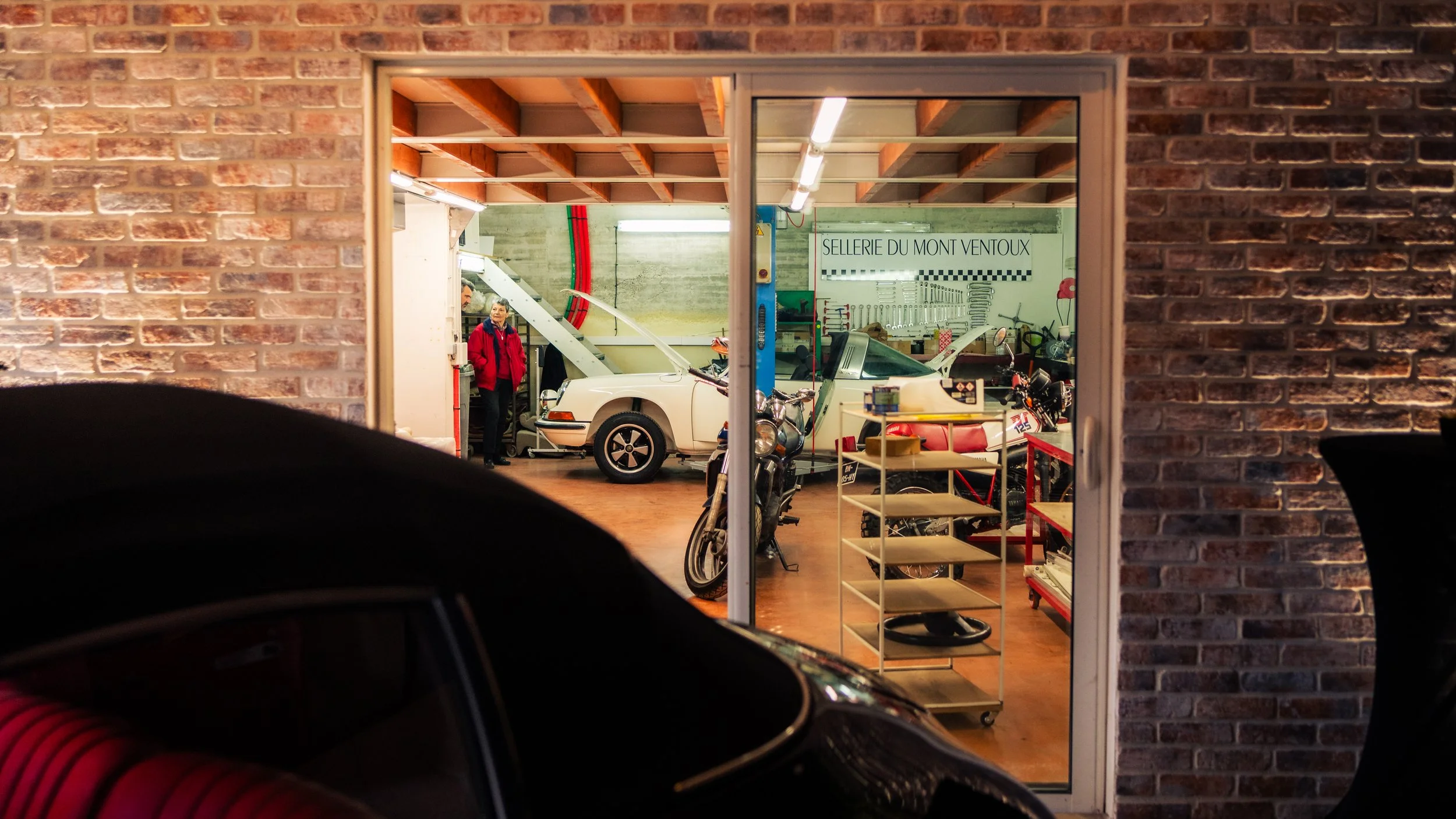 View through a glass window into a workshop with a classic white Porsche with its hood open, surrounded by motorcycles, tools, and shelves, with a person in a red jacket inside.