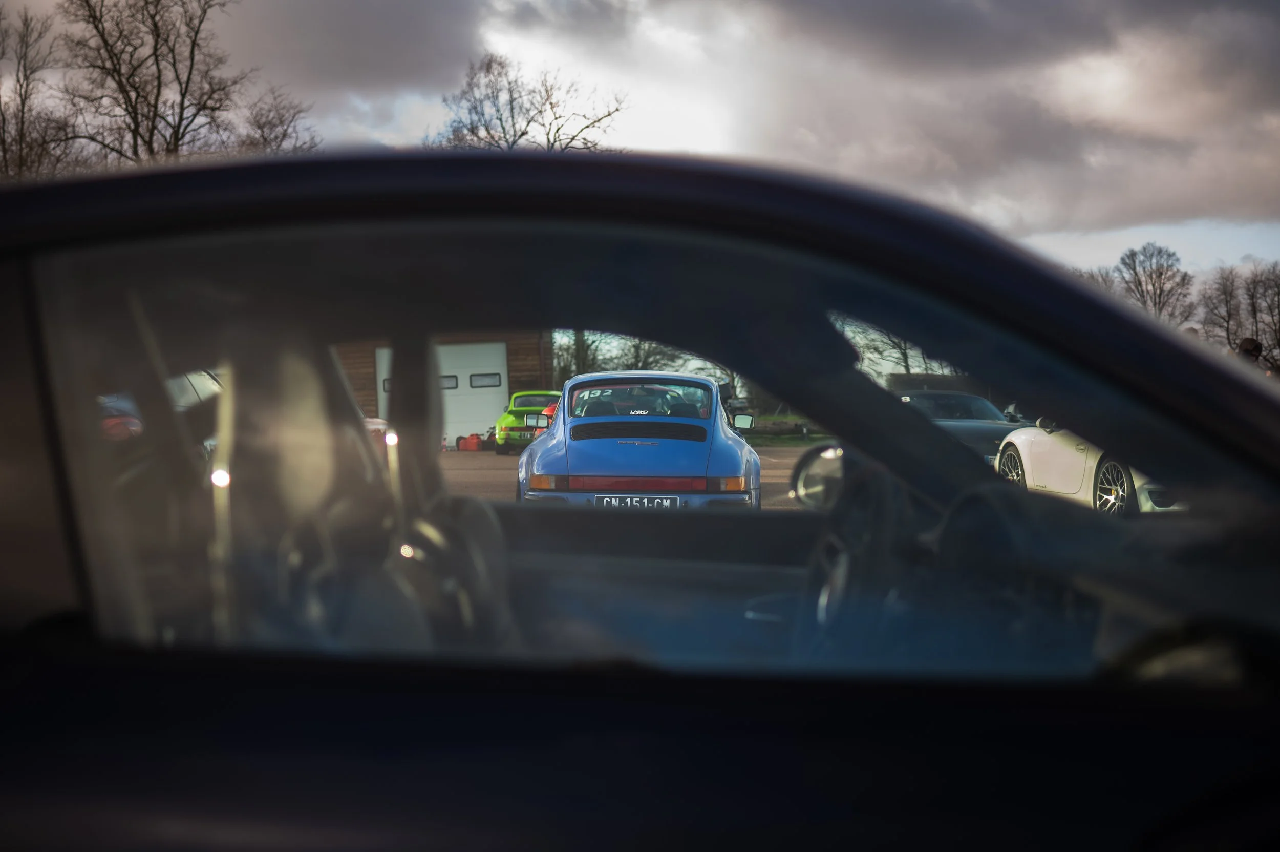 View from inside a car looking out through the rear window at a parking lot of classic and sports cars, including a blue Porsche in the center, with other cars and a garage in the background under a cloudy sky.