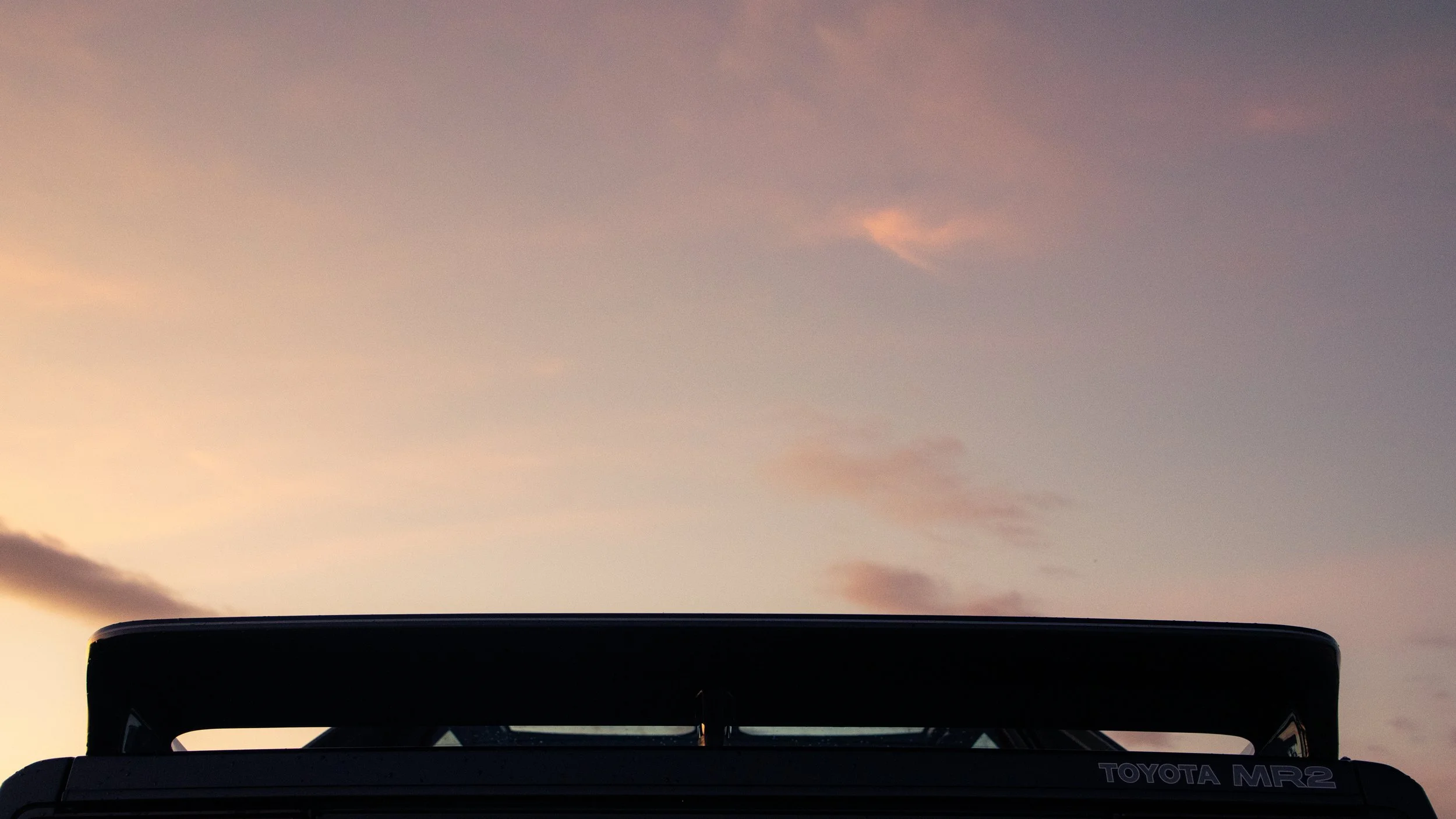The image shows the rear end of a Toyota MR2 car with a spoiler, set against a colorful evening sky with pink and purple clouds.