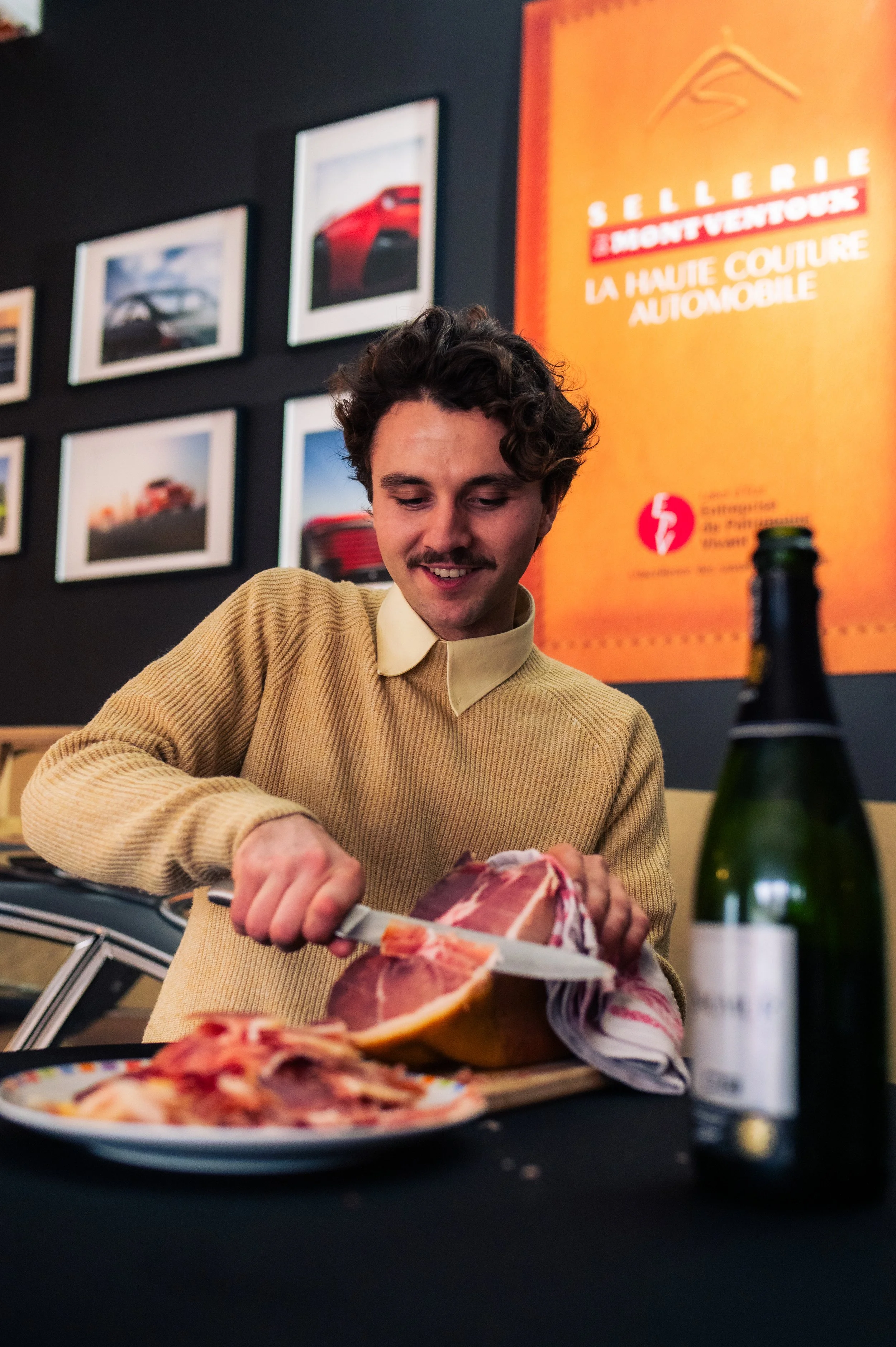A man with curly hair and a mustache slicing a large piece of raw meat on a cutting board in a dining setting with plates of meat, a beer bottle, and framed pictures of cars on the wall behind him.