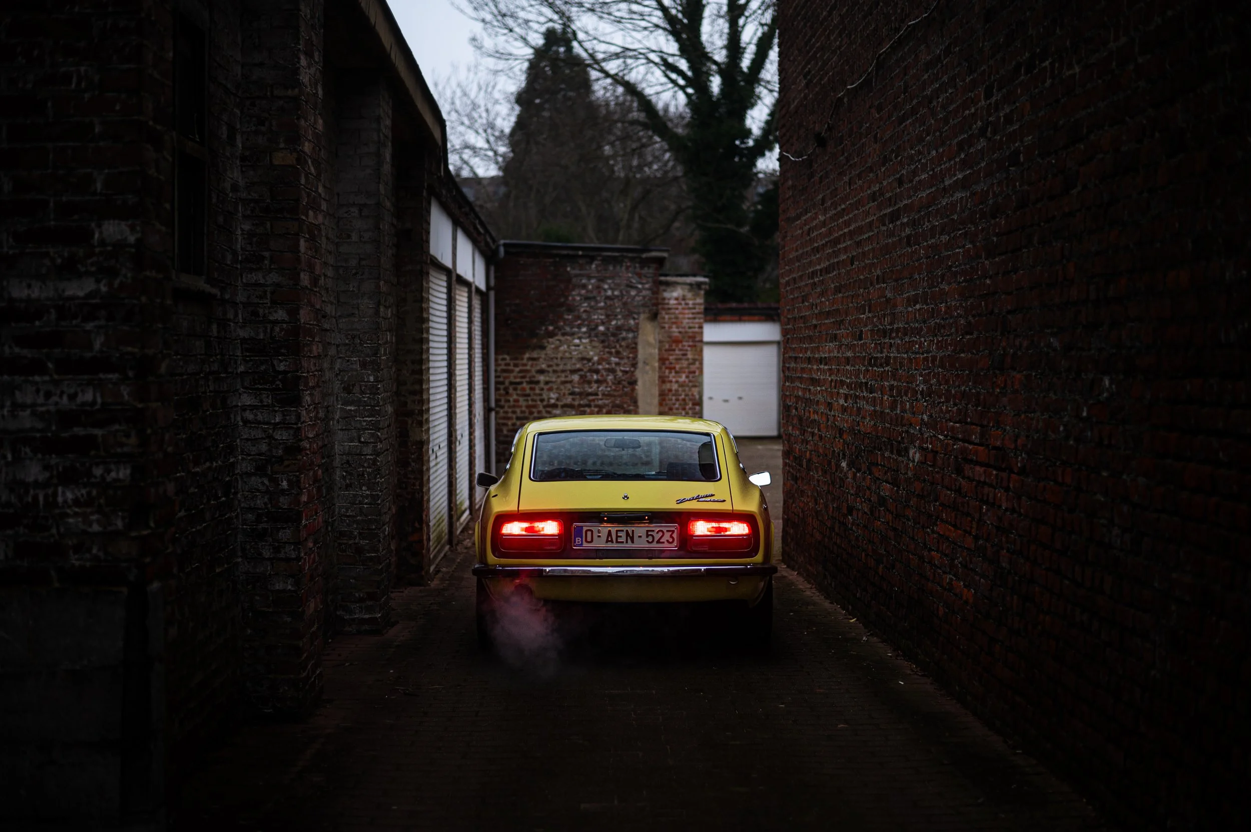 A yellow Datsun 240Z parked in a narrow alley between brick buildings, with its rear lights on and visible exhaust fumes.