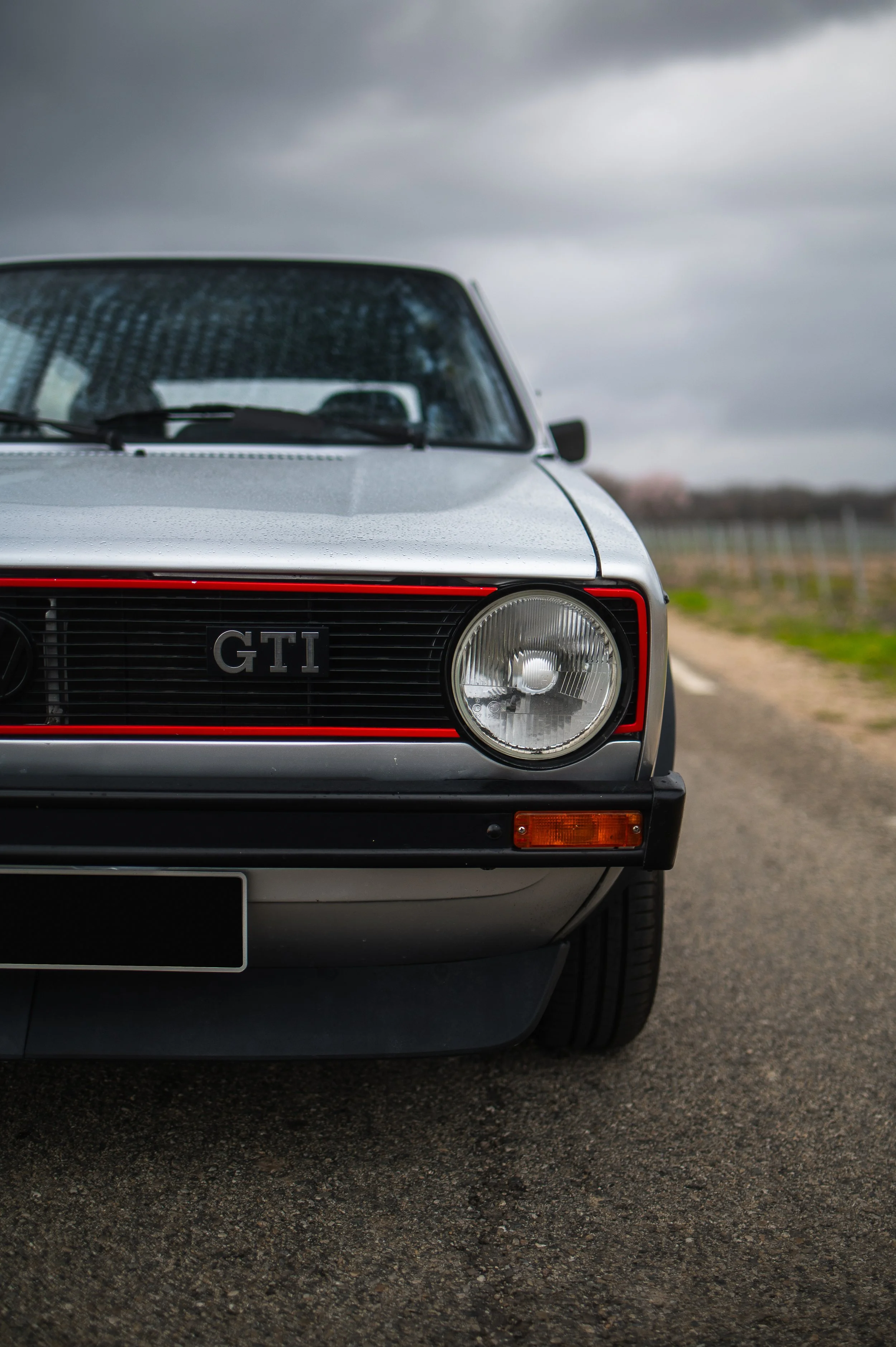 Close-up of a classic gray vintage car with a black grille and round headlight, parked on a rural road with overcast sky.