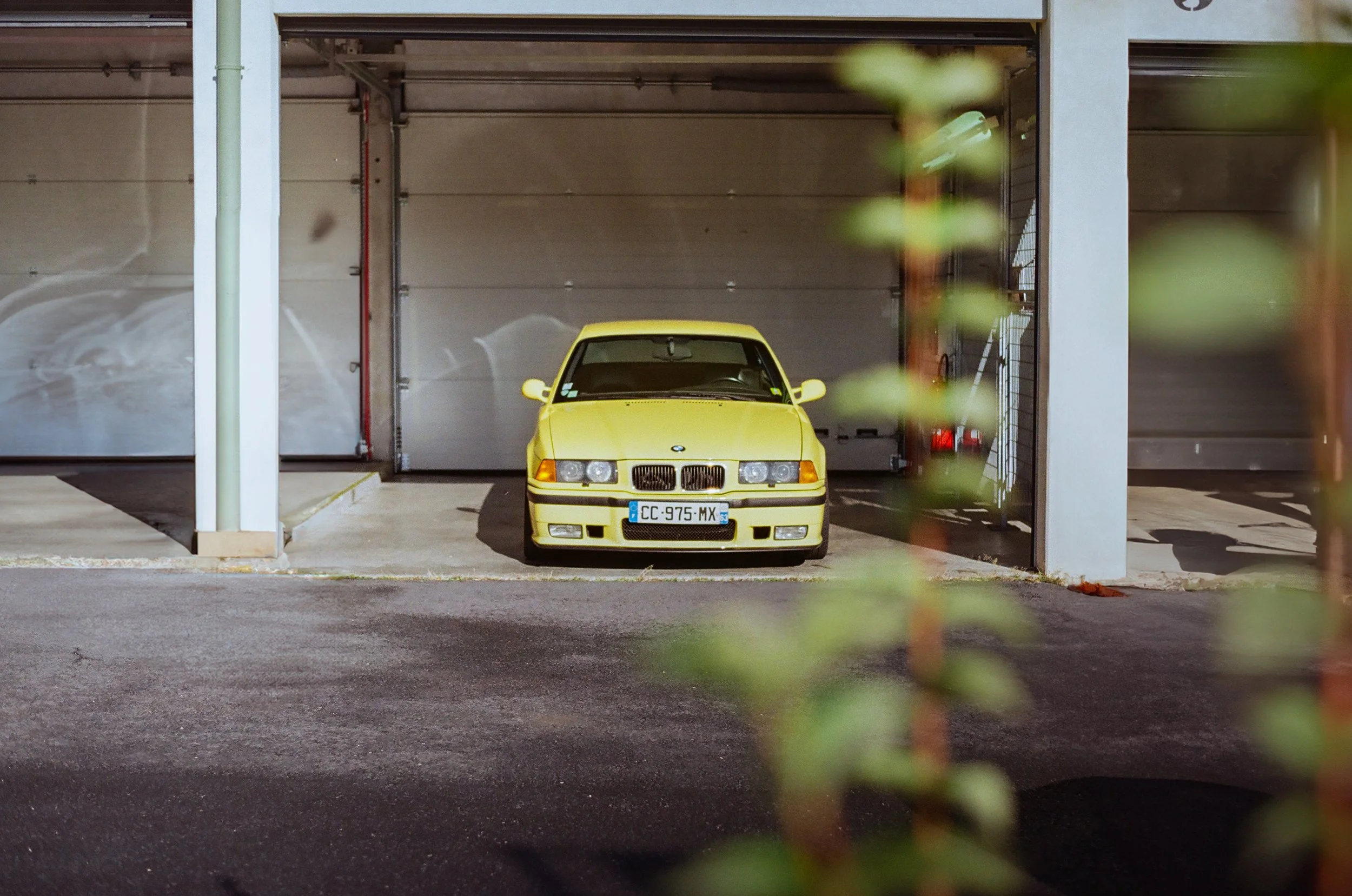 A yellow BMW M3 E36 car parked in a garage at Charade racetrack with a closed gray door, partially obscured by green out-of-focus leaves in the foreground.