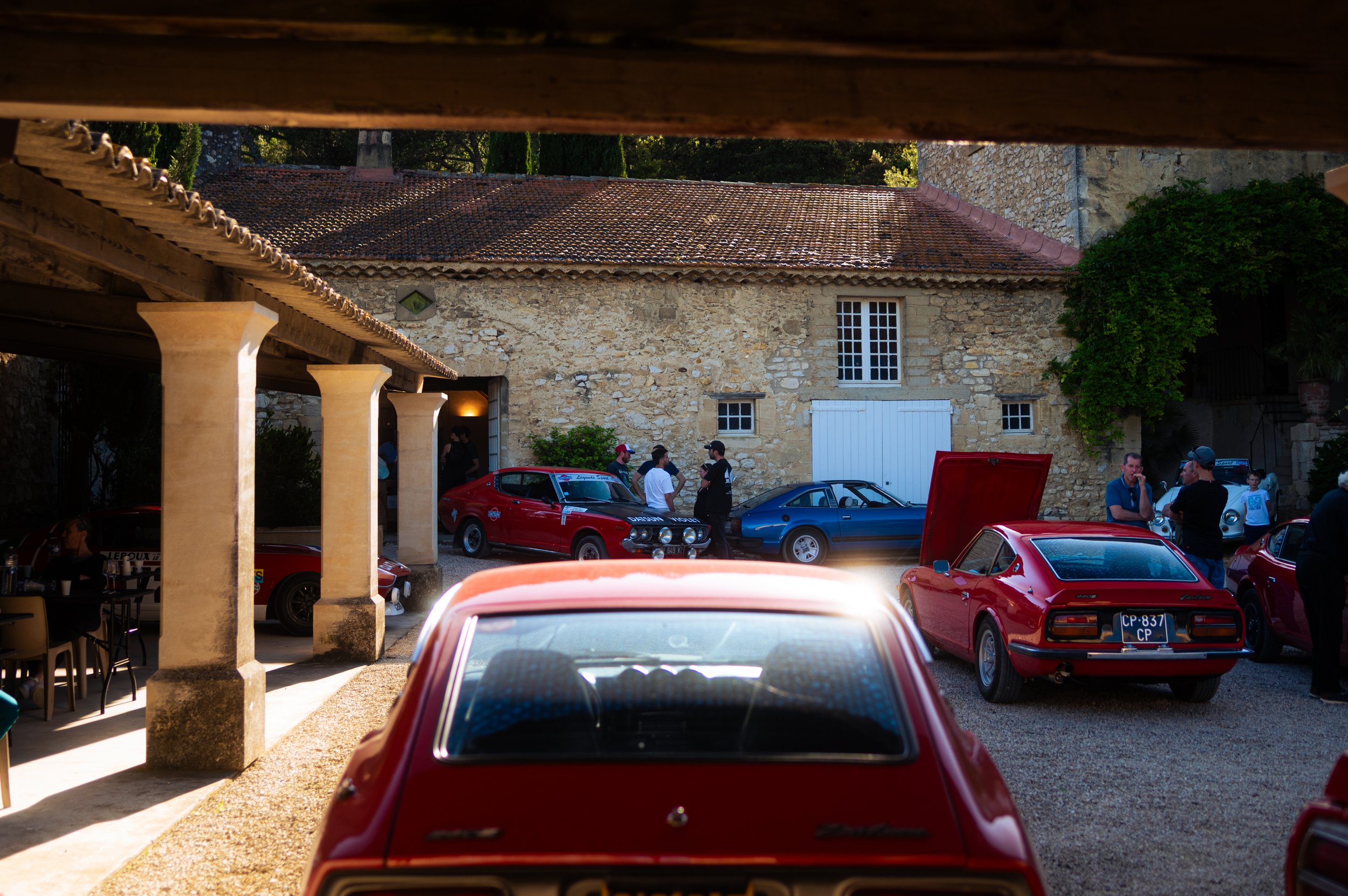 Several classic sports cars, predominantly red and blue, parked in a courtyard with people socializing nearby. The scene is captured in daylight with a stone building and arches in the background.