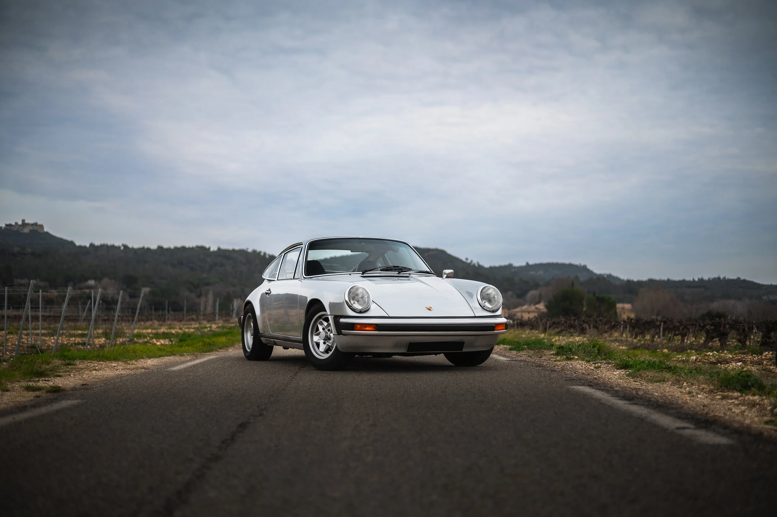 A classic silver Porsche 911 parked on a rural road with mountains and cloudy sky in the background.