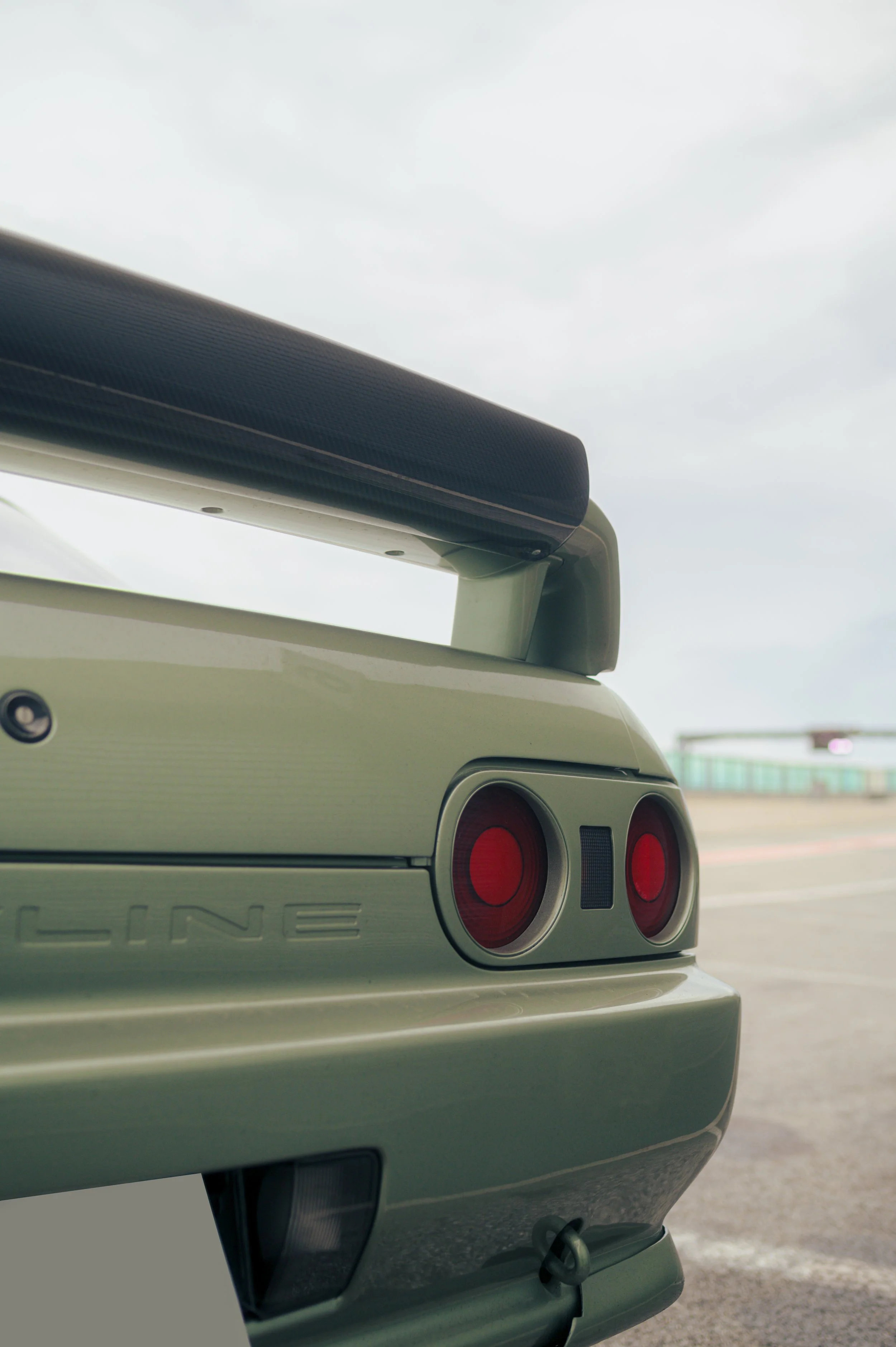 Close-up of the rear of a Nissan Skyline R32 in Millenium Jade, showing red tail lights and a large rear spoiler on a cloudy day.