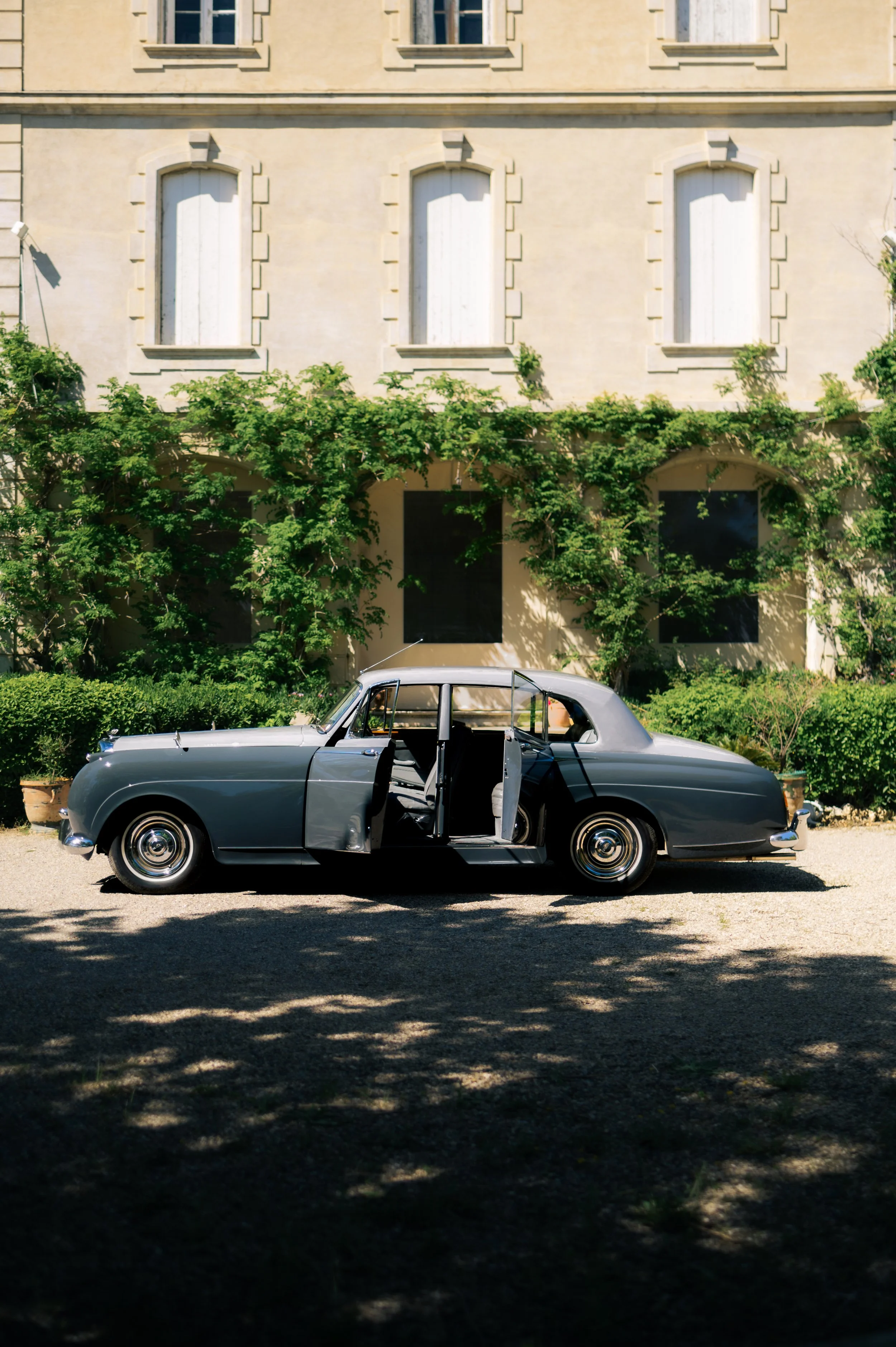 A 1958 dual-tone gray Bentley Mulliner S1 parked in front of a beige building with green foliage, and the car's passenger doors open.