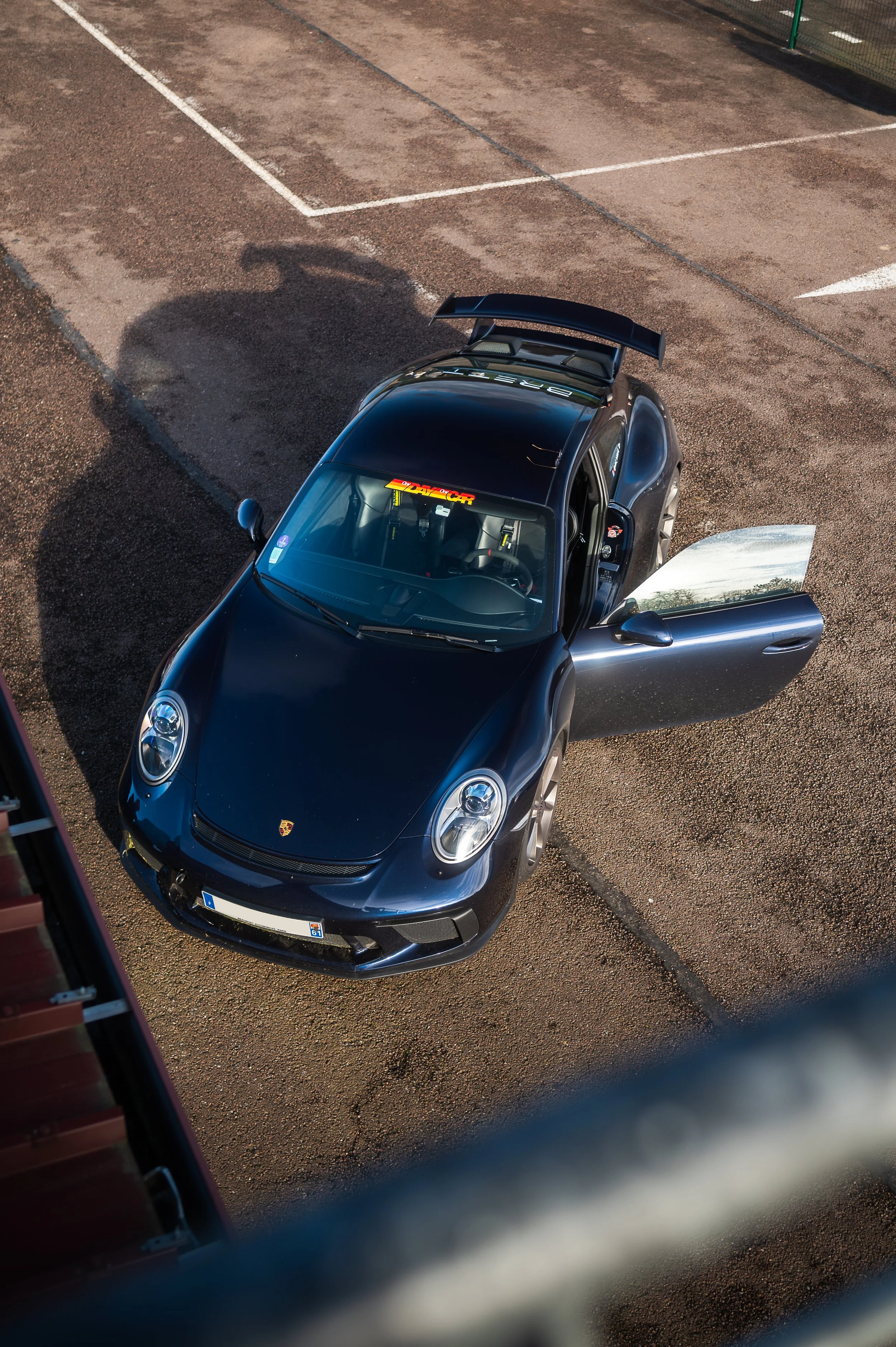 Blue Porsche 911 (991.2) GT3 with open door parked in the paddock of Circuit des Ecuyers, captured from an overhead angle.