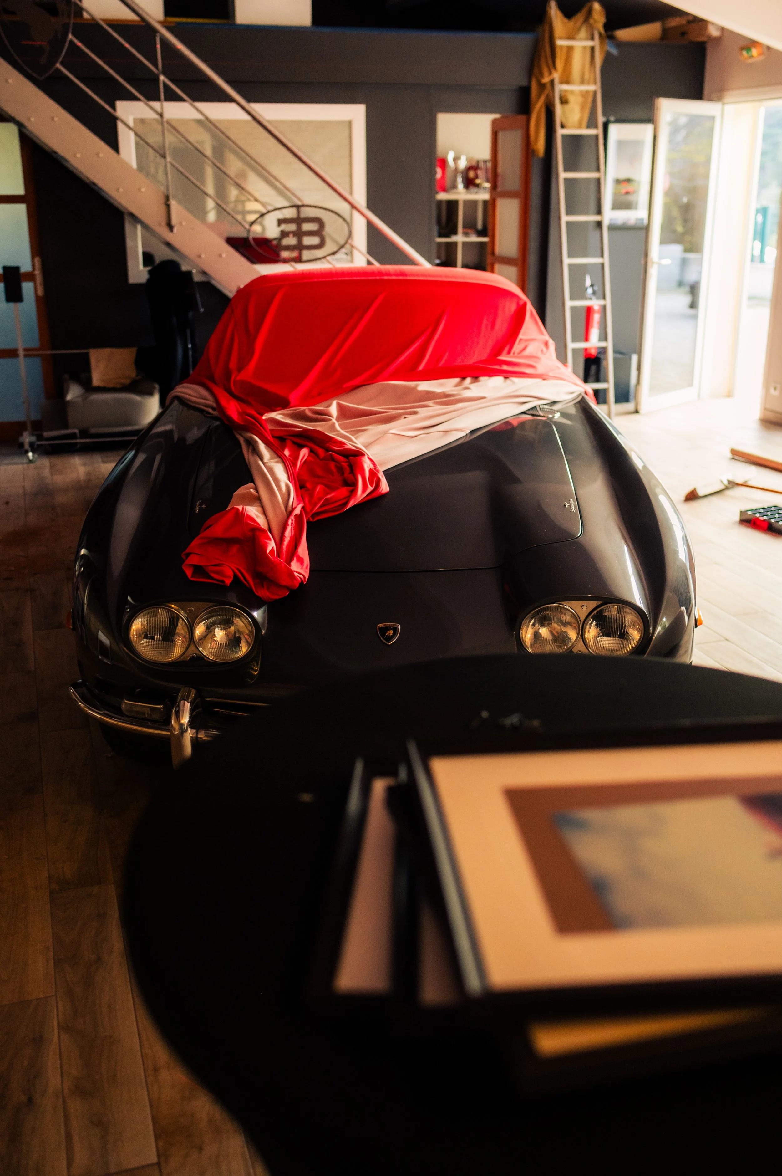 A black vintage Lamborghini with a red cover partially removed, inside a garage or showroom, with a ladder, shelves, and a sliding glass door in the background.