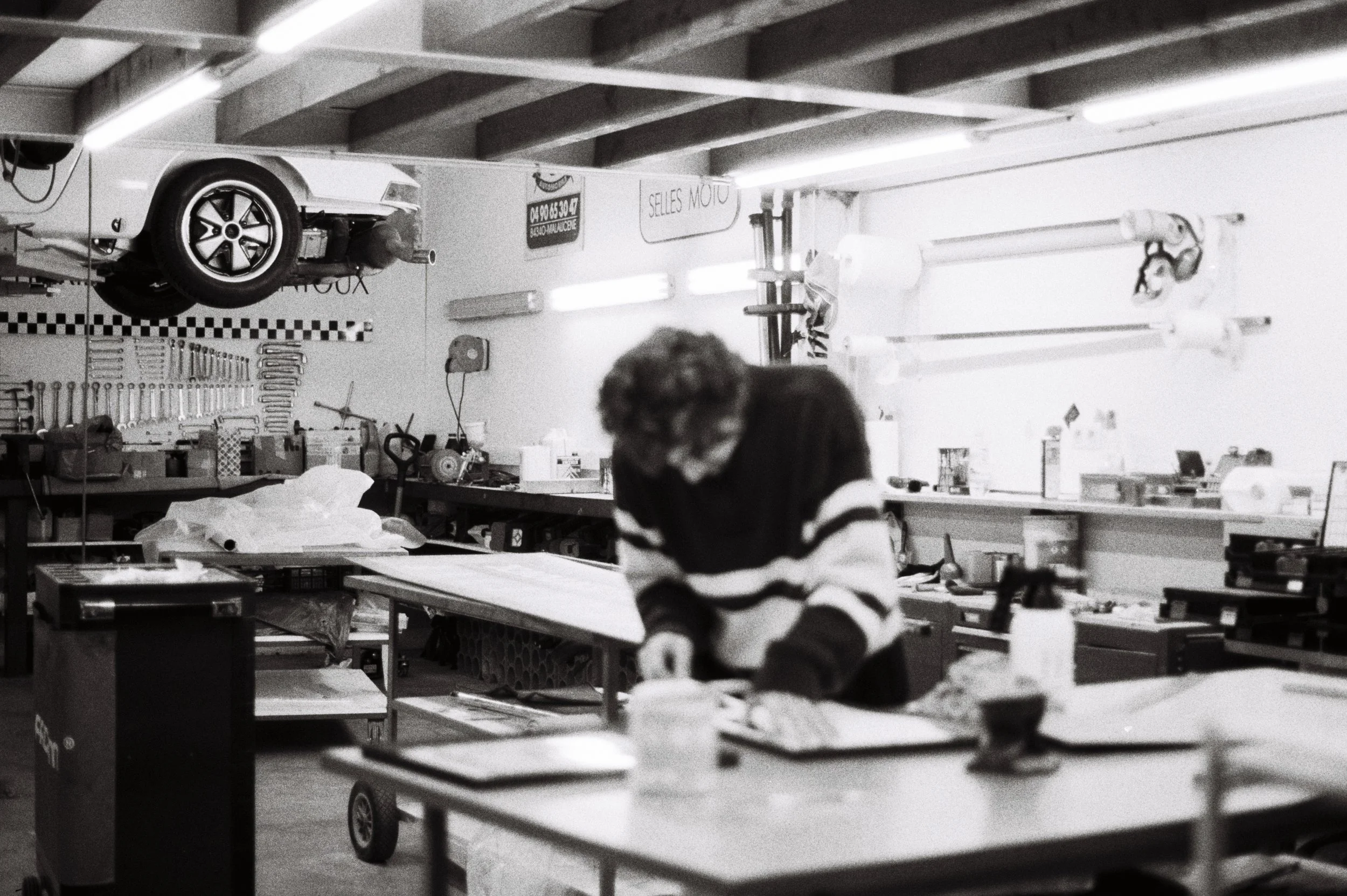 A person working leather at a cluttered workbench in a garage or workshop, with a car mounted on a lift above and tools on the wall.