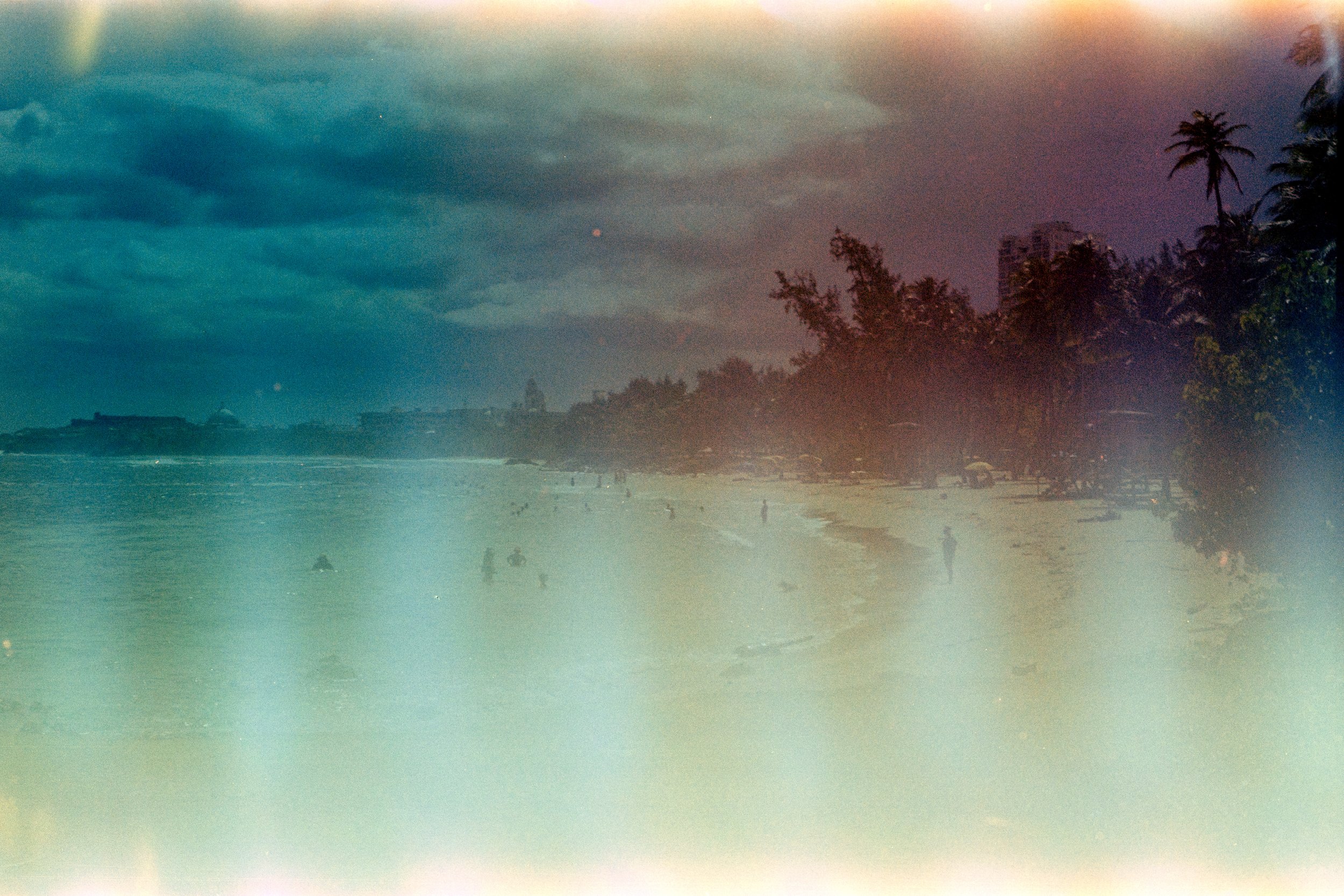 A beach with people swimming and walking along the shore, palm trees, buildings in the background, a cloudy sky.