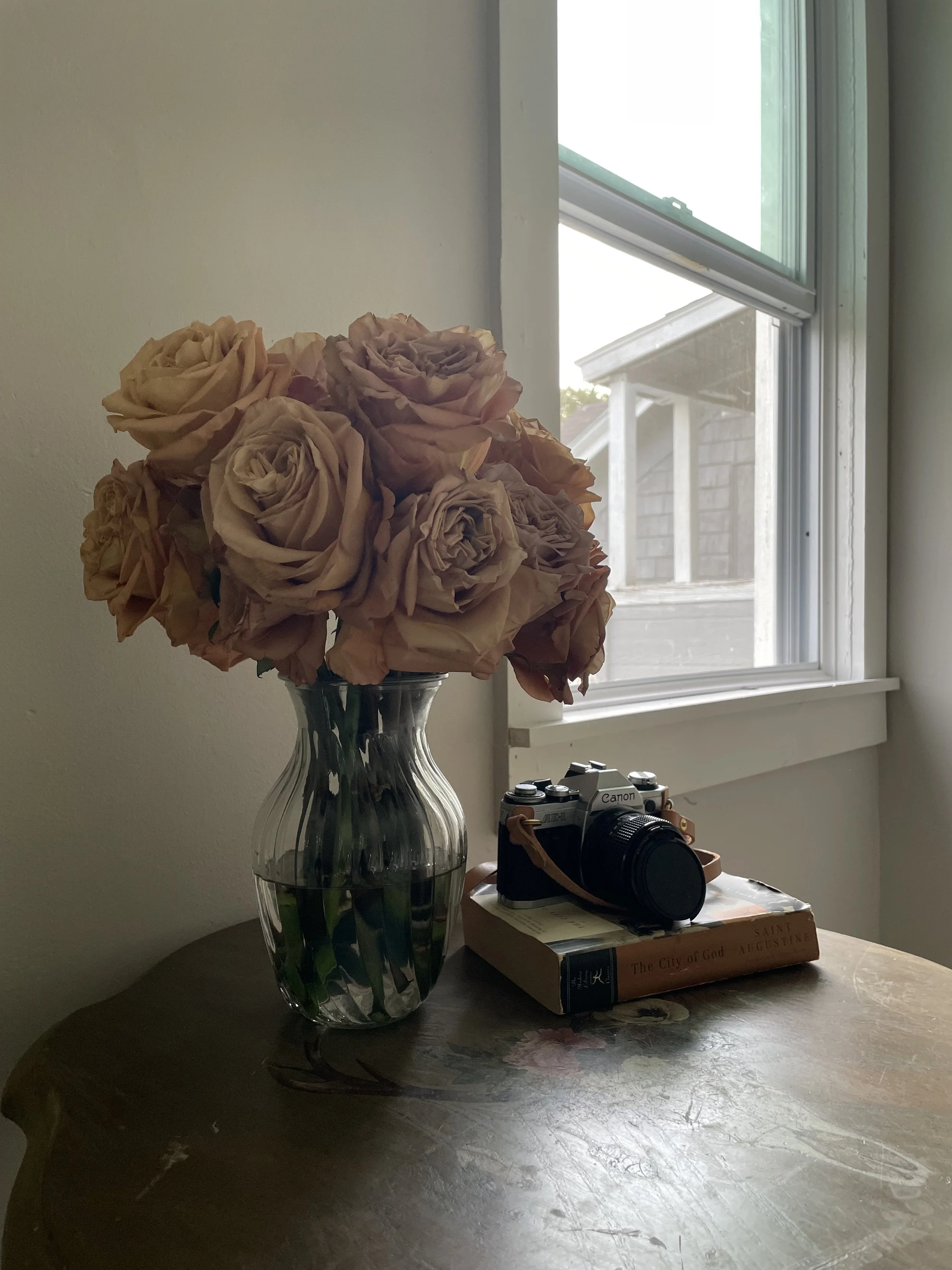 A clear glass vase with beige roses on a wooden table, next to a vintage Canon camera placed on a book titled 'The City of God' by Saint Augustine near a window.