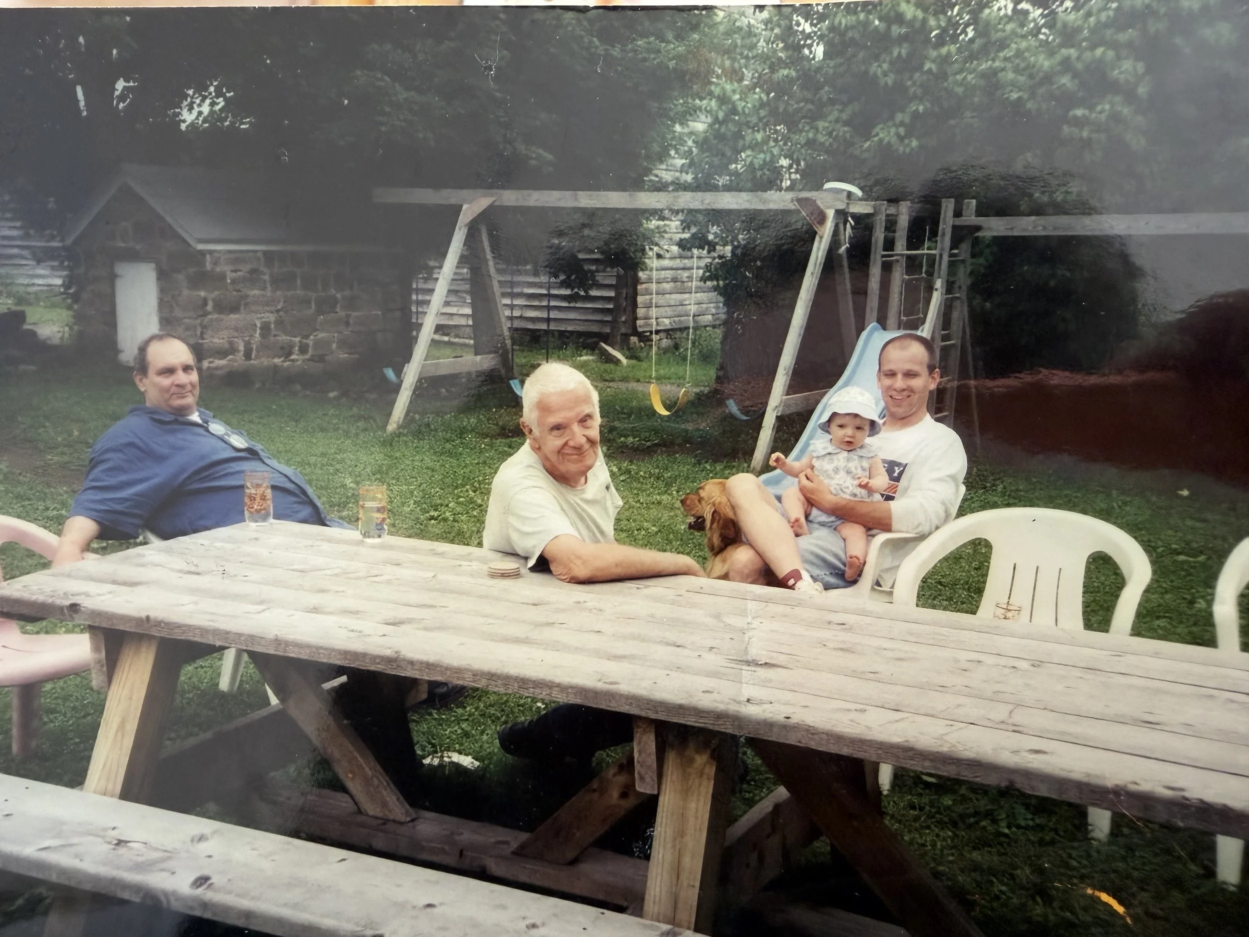 A family of four and a dog sitting around a wooden picnic table in a backyard, with a swing set and small brick building in the background.
