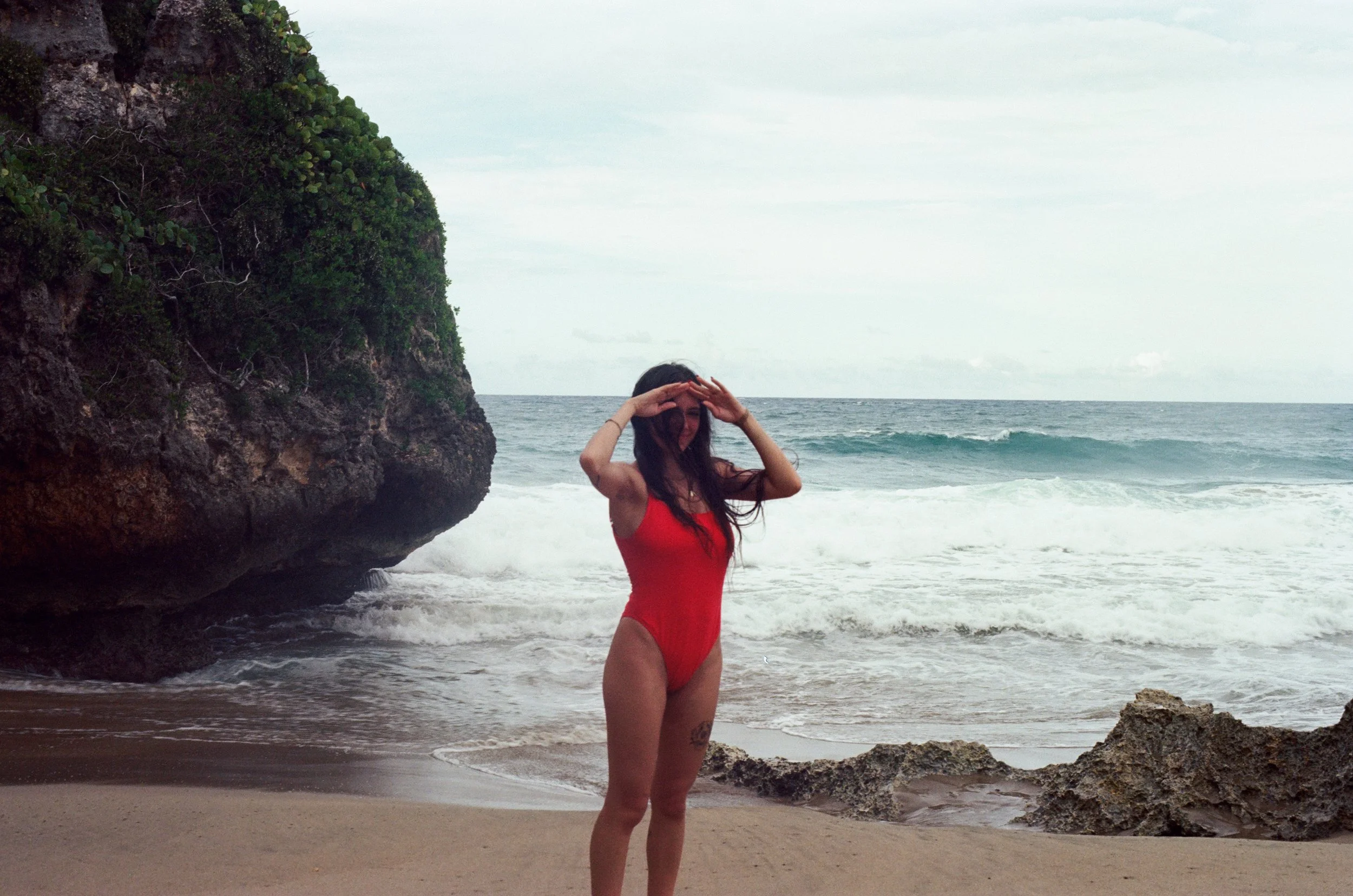 A woman in a red swimsuit standing on a sandy beach near rocky formations with the ocean in the background, shielding her eyes from the sun.