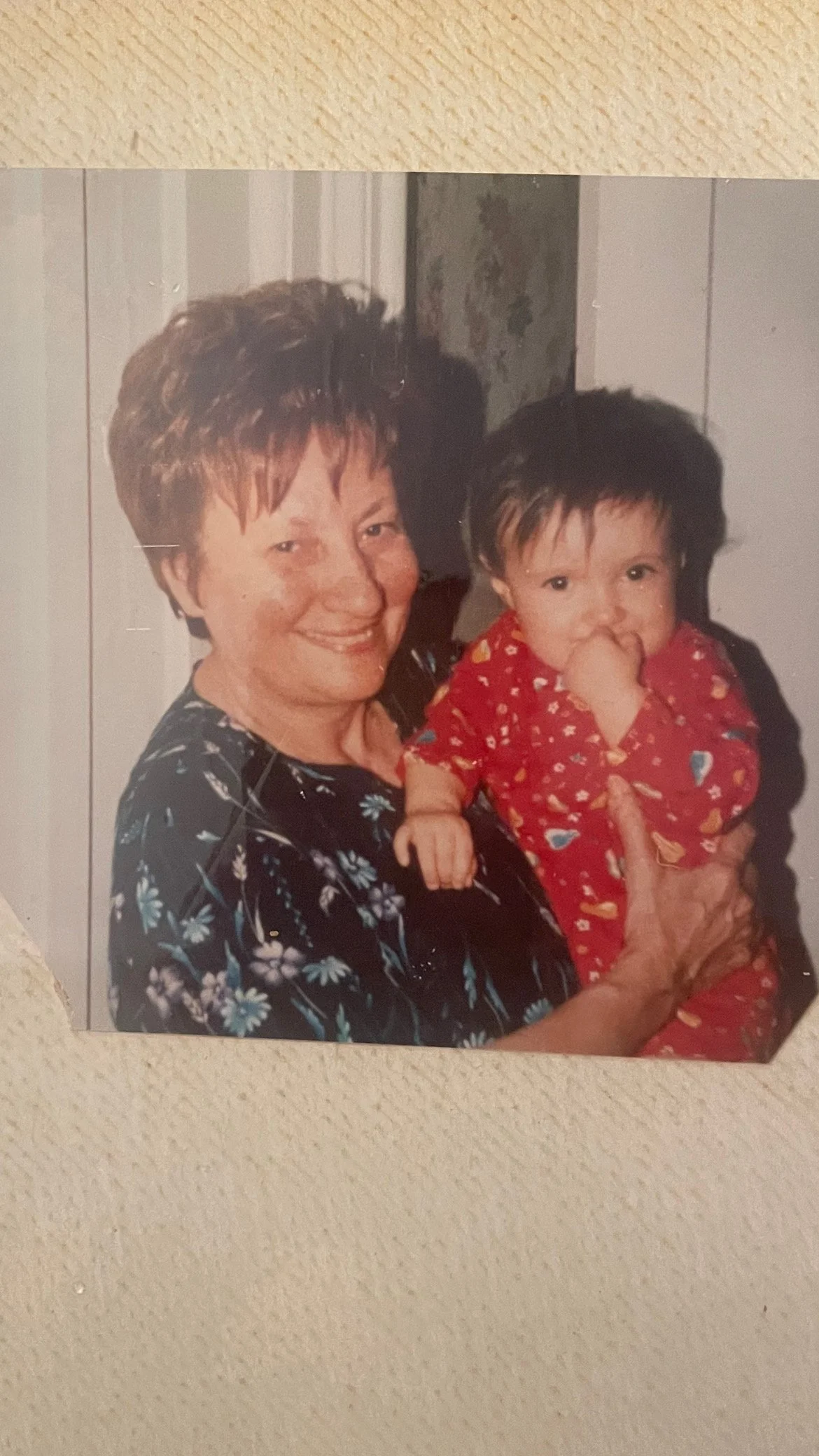 An elderly woman smiling and holding a young child who is wearing a red pajamas with colorful patterns, the woman is wearing a dark floral blouse, and they are indoors near a wall.