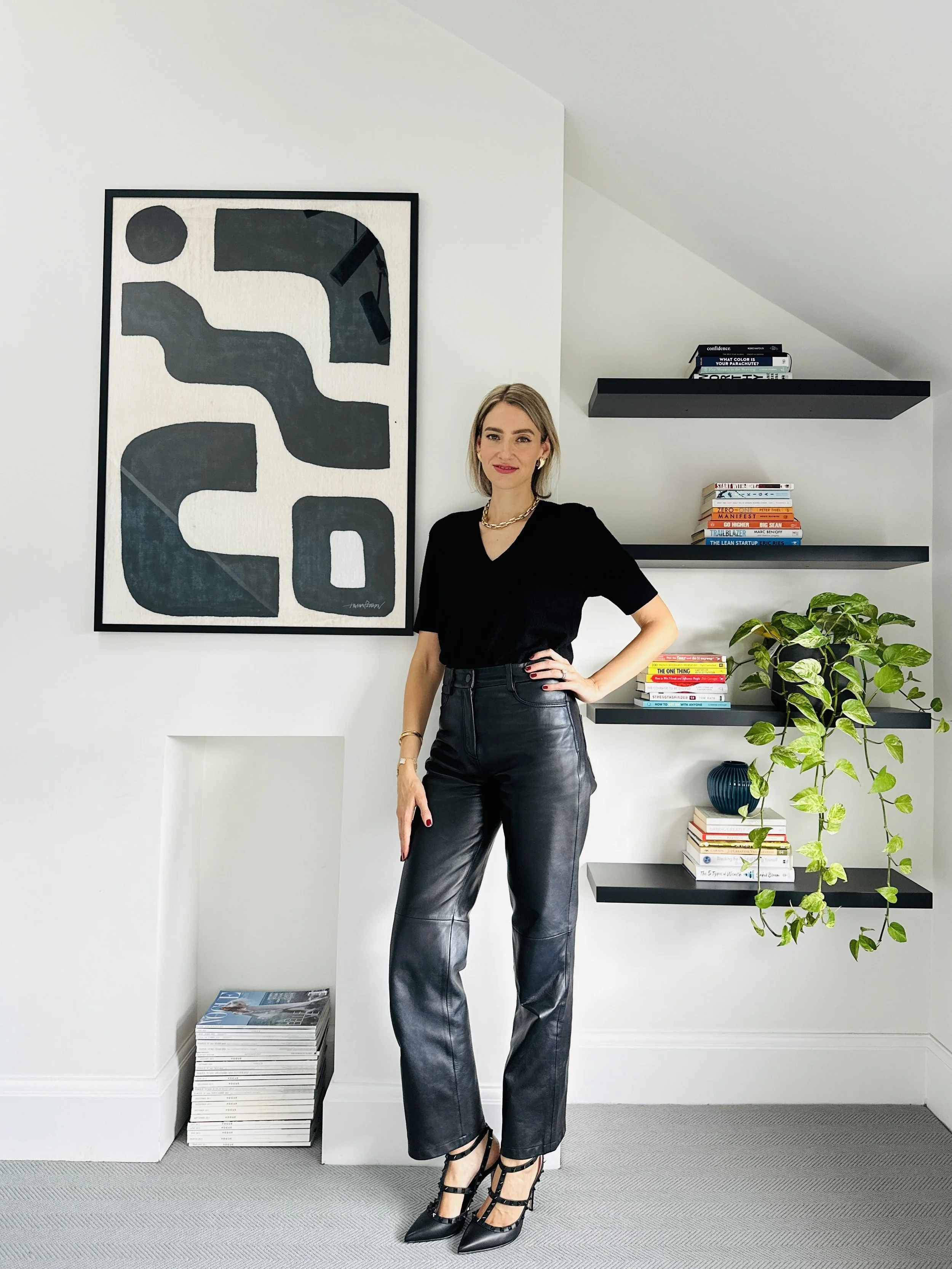 A woman with blonde hair and red lipstick standing in a modern white room, wearing black leather pants and a black top, with one hand on her hip, next to a wall with black floating shelves filled with books and a potted plant.