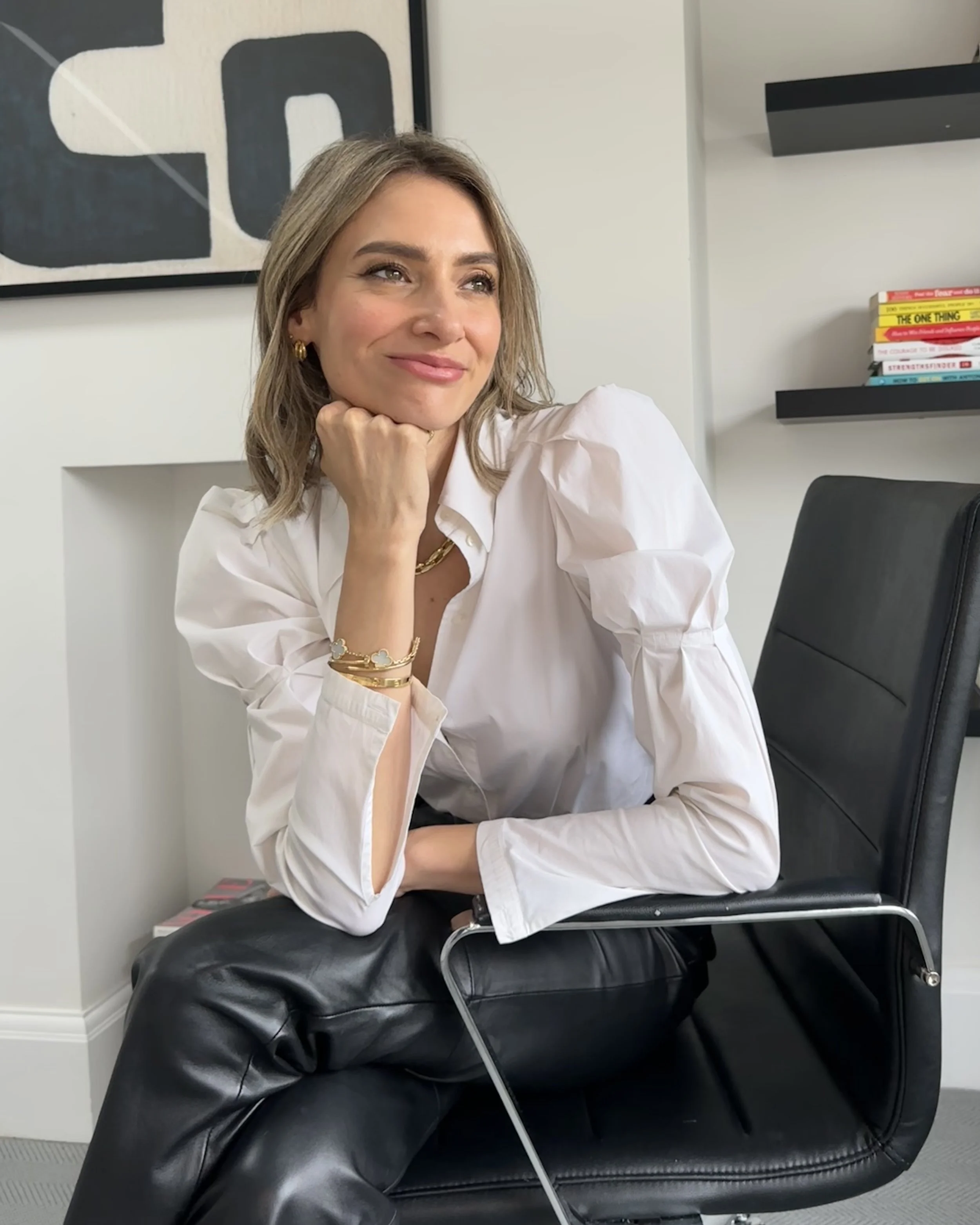 A woman with shoulder-length blonde hair sitting in a black leather office chair, smiling with her chin resting on her hand, wearing a white blouse with puffy sleeves, gold hoop earrings, layered gold bracelets, and a gold chain necklace, in a modern office space with abstract wall art and books on black floating shelves.