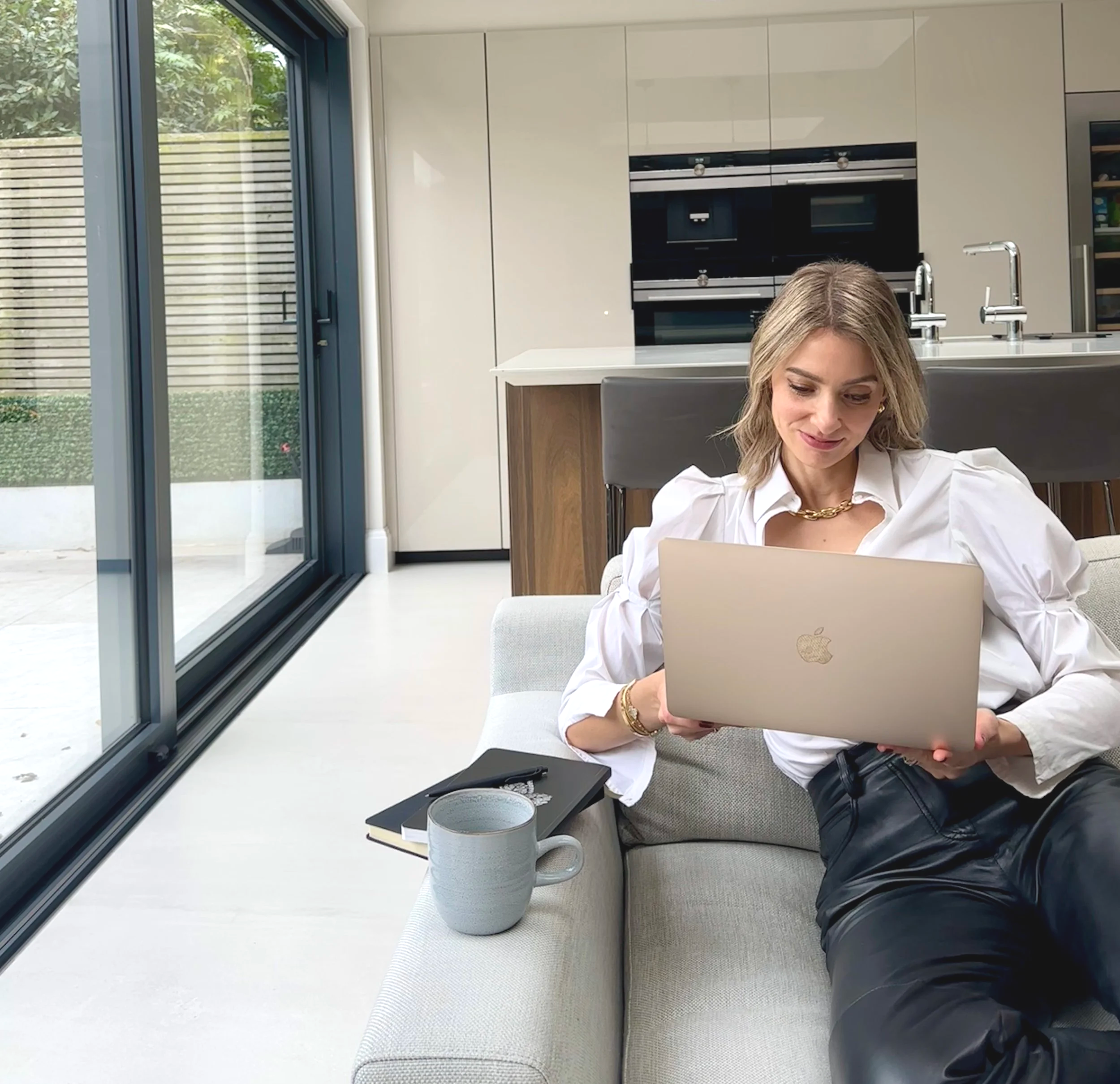 A woman sitting on a light-colored sofa in a modern kitchen, looking at her laptop. There is a coffee mug and a notebook with a pen on the sofa armrest beside her. Large glass doors lead to an outdoor space.