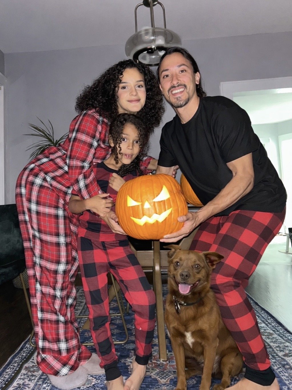 A family of three and a dog celebrating Halloween indoors. They are wearing matching red and black plaid pajamas. They hold a carved pumpkin lantern with a spooky face. The children and the dog look happy, and the adults smile.