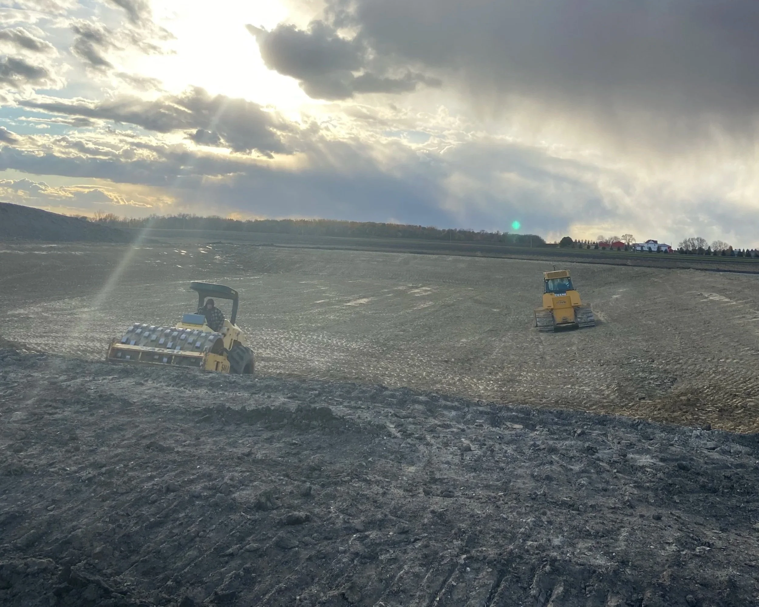 Two workers operating small yellow construction vehicles on a large plowed field under a cloudy sky with the sun peeking through.