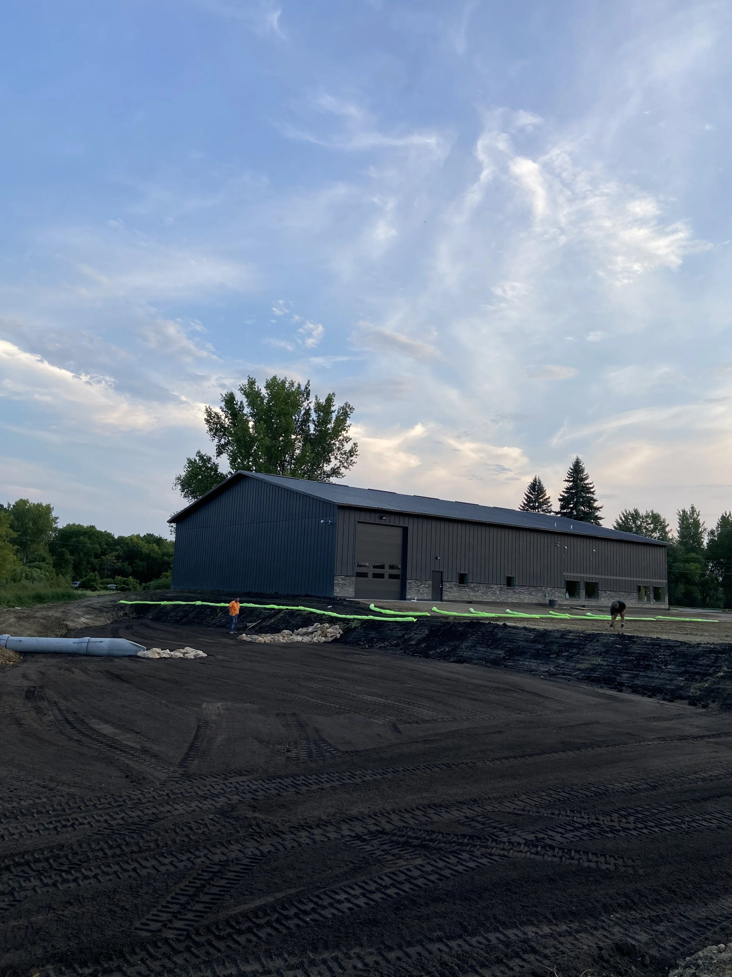 Construction site with a large blue industrial building and workers installing drainage pipes, with a dirt ground, some greenery, and a partly cloudy sky.
