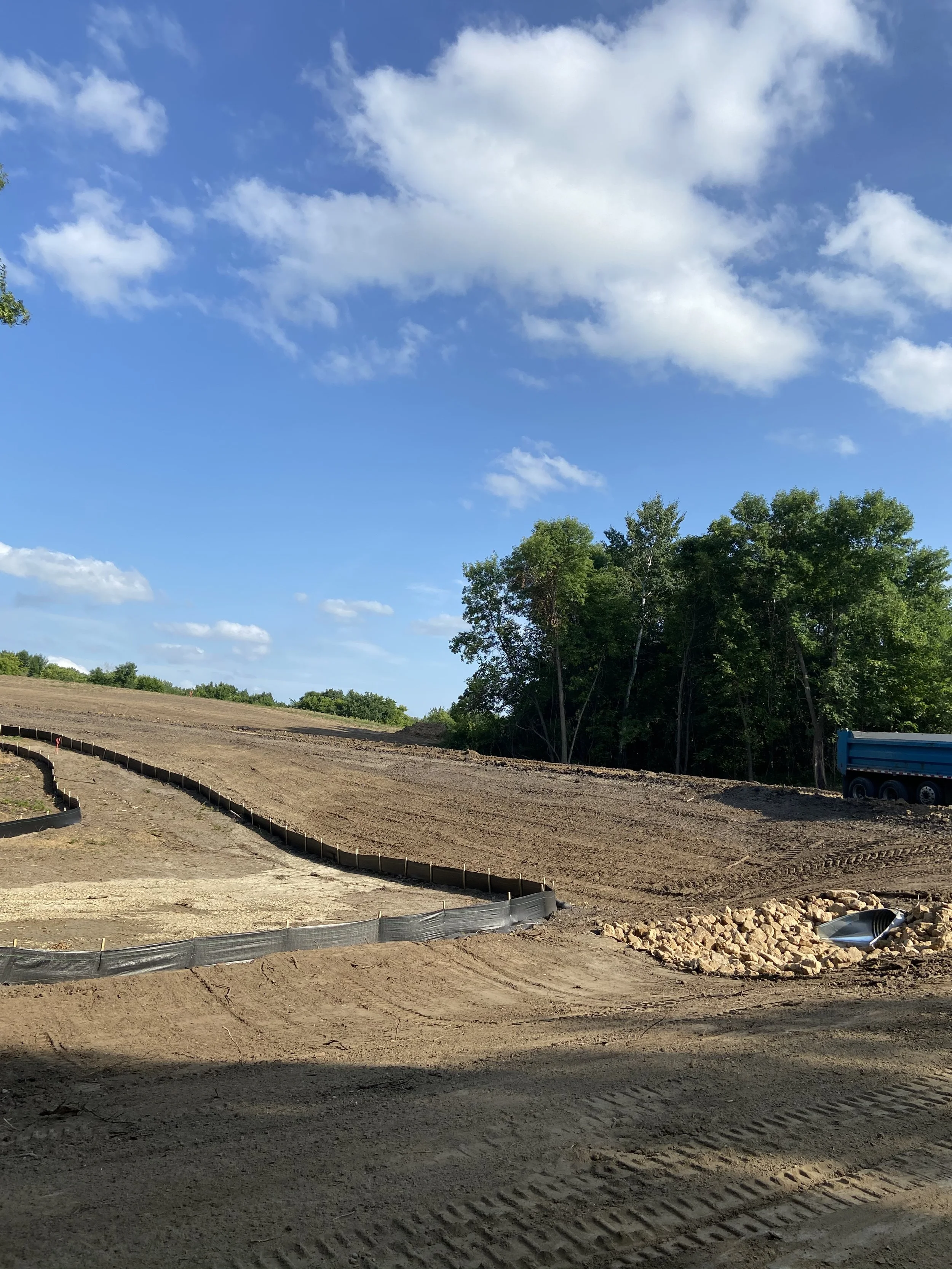 Construction site with dirt, rocks, a blue truck, green trees, and a partly cloudy sky.