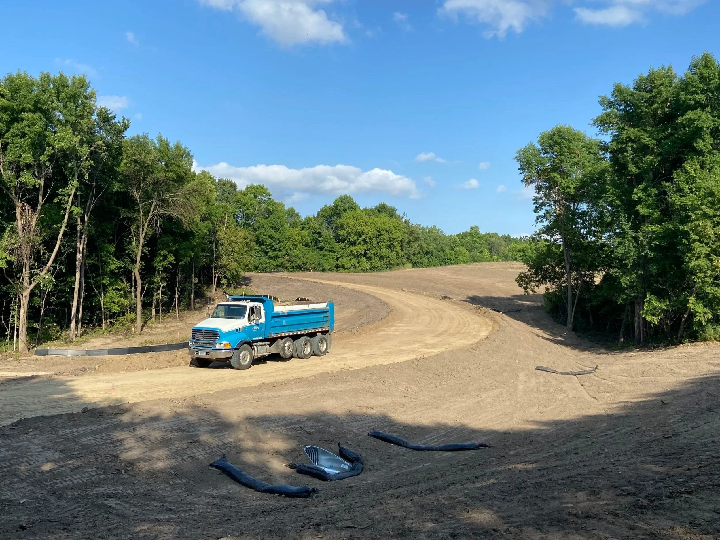 A dirt road winding through a construction site surrounded by green trees with a blue dump truck parked on the dirt pathway. Construction materials and debris are scattered on the ground, under a partly cloudy sky.