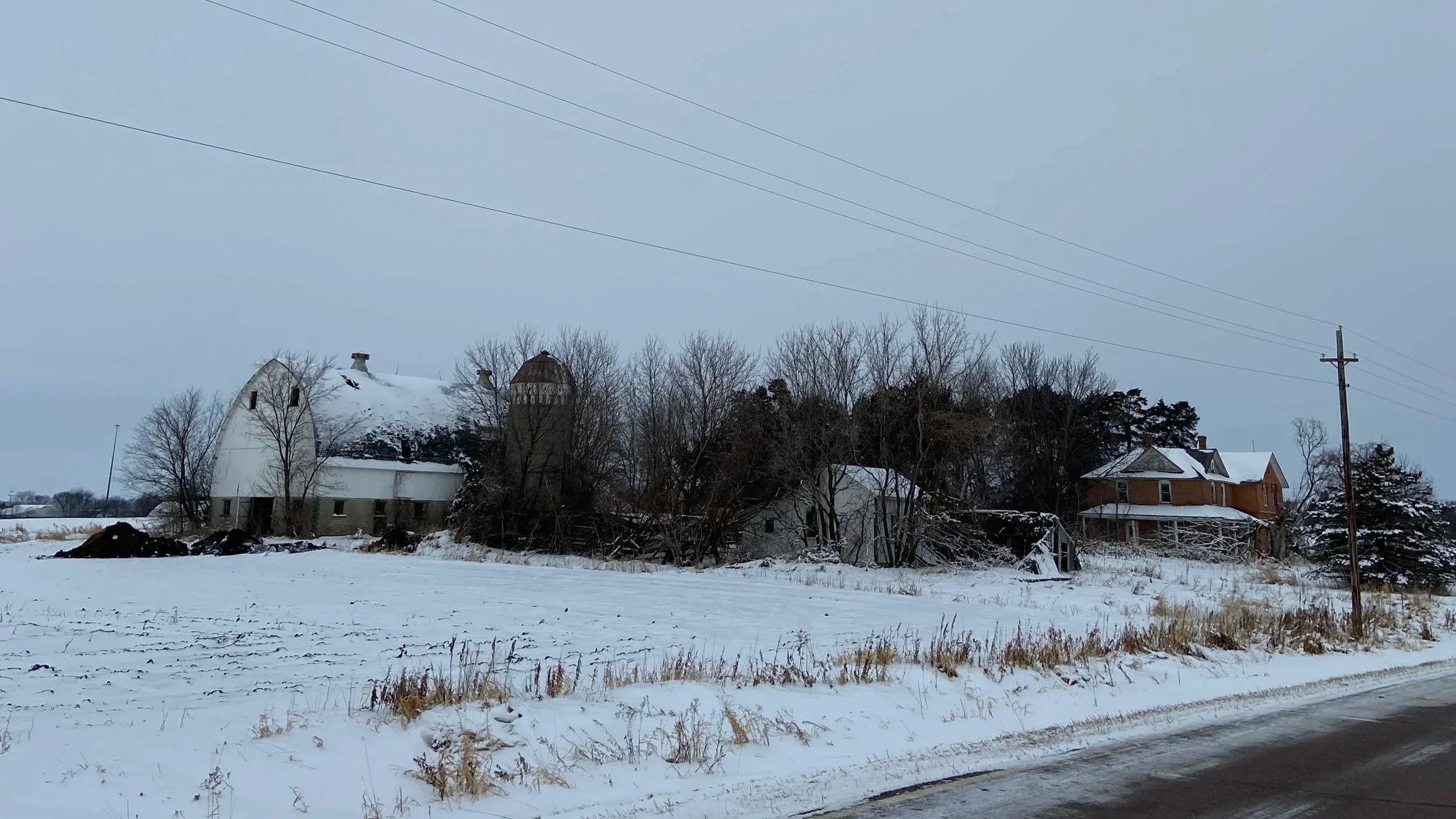 Snow-covered farm buildings and house in a rural winter landscape with leafless trees, power lines, and a snowy field in the foreground.