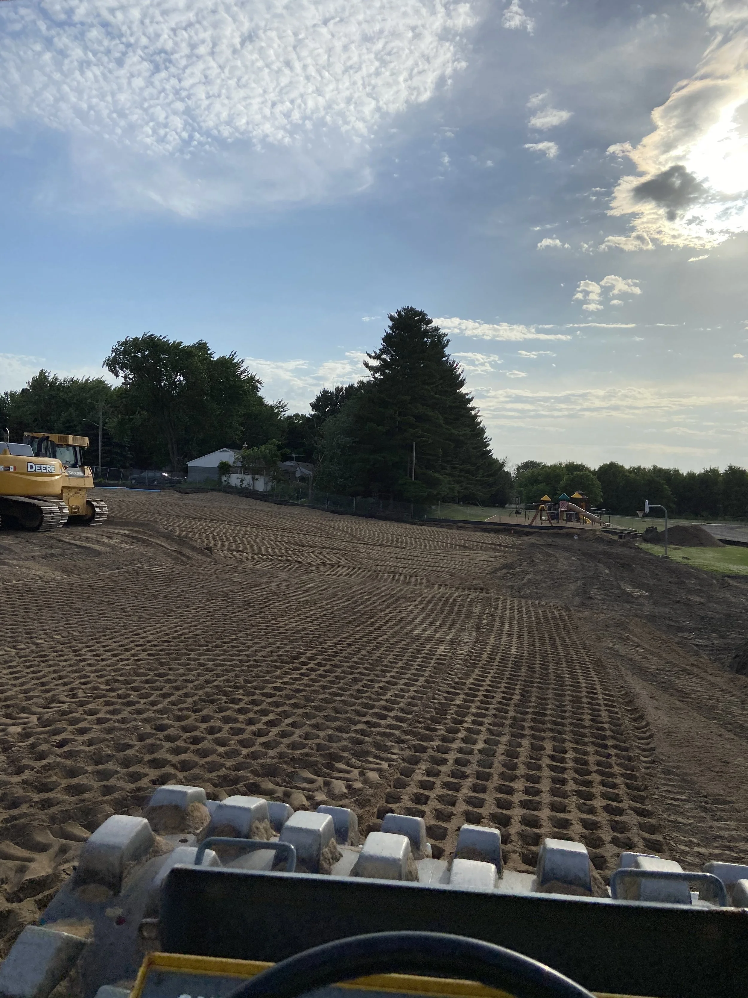 View from a construction vehicle showing a dirt field being worked on, with a yellow bulldozer on the left, some trees, a playground in the distance, and a partly cloudy sky overhead with the sun shining through the clouds.