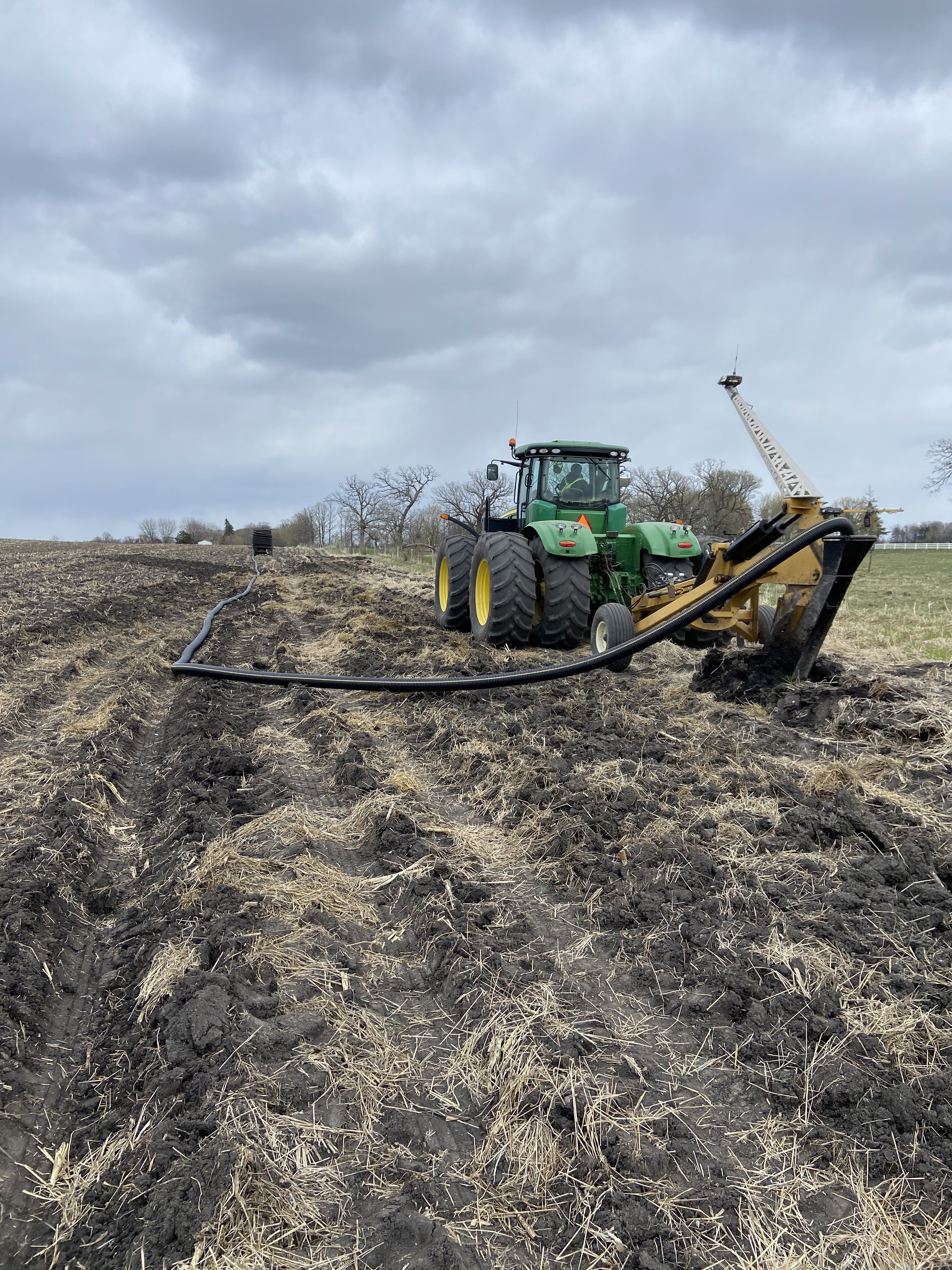 A tractor working on tiling a field under a cloudy sky, with a long hose connected to a piece of farm equipment.