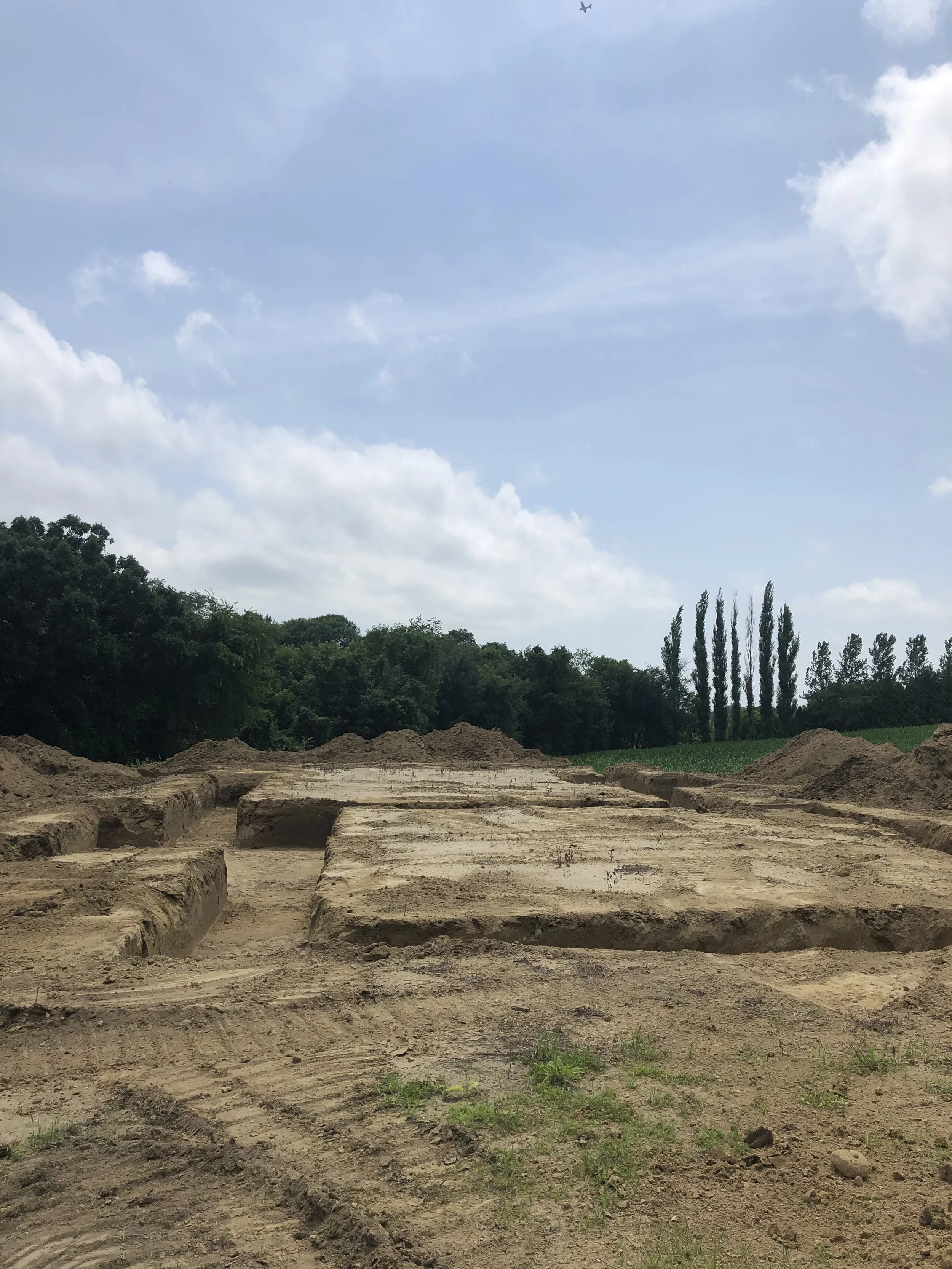 Excavated land with clear rectangular trenches, surrounded by trees and a partly cloudy sky.
