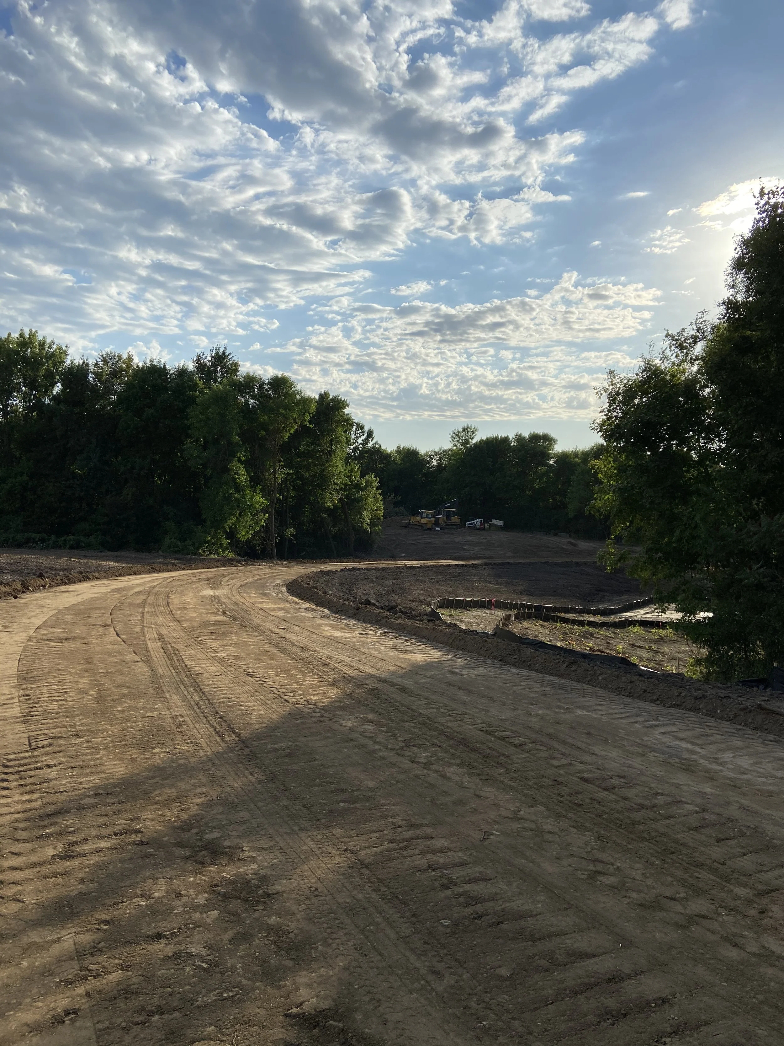 A dirt road on a construction site with trees on both sides and a sky with scattered clouds.