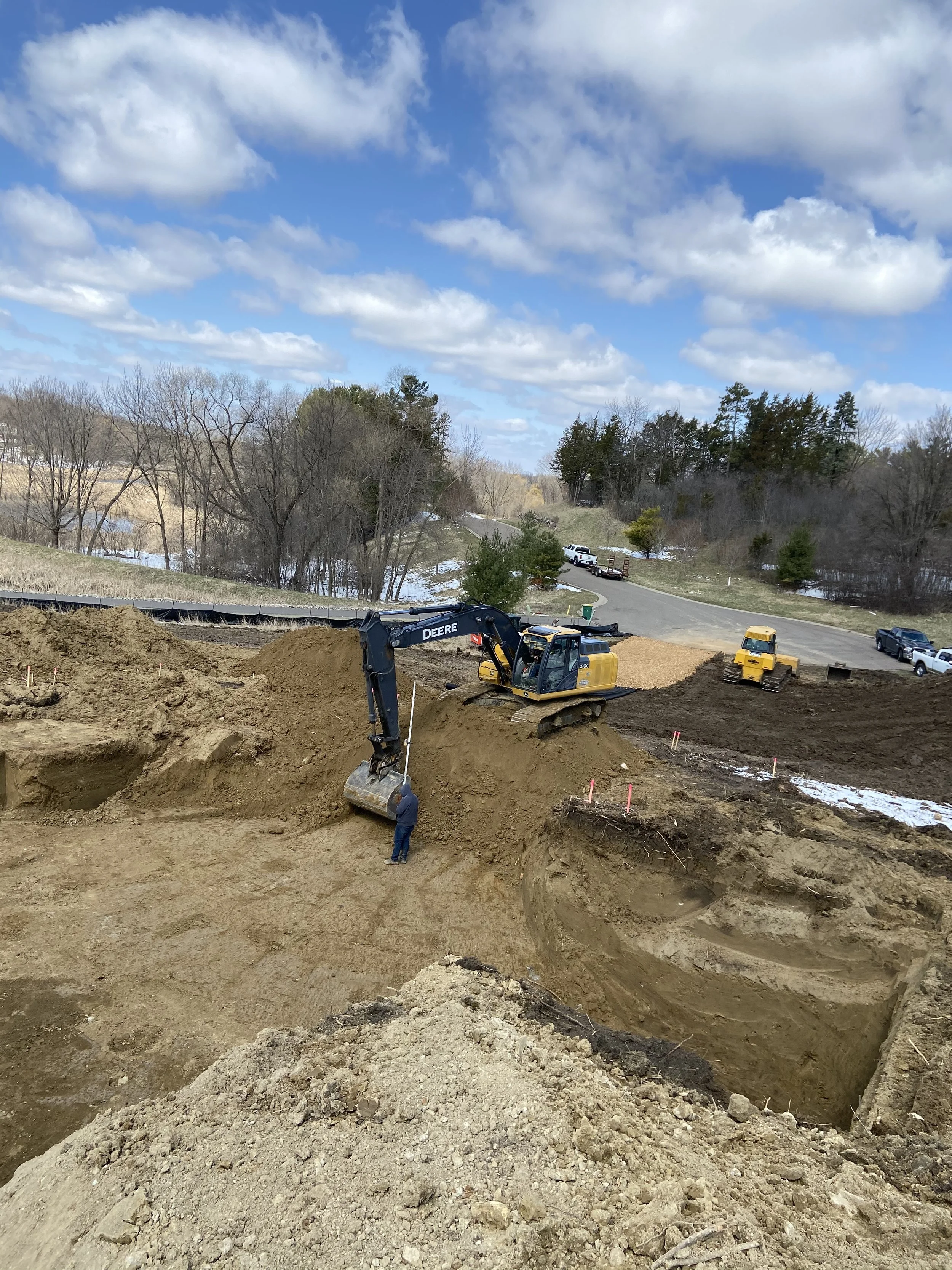 Construction site with a large excavator digging a deep hole in the ground, and a worker standing nearby, during daytime with partly cloudy sky.