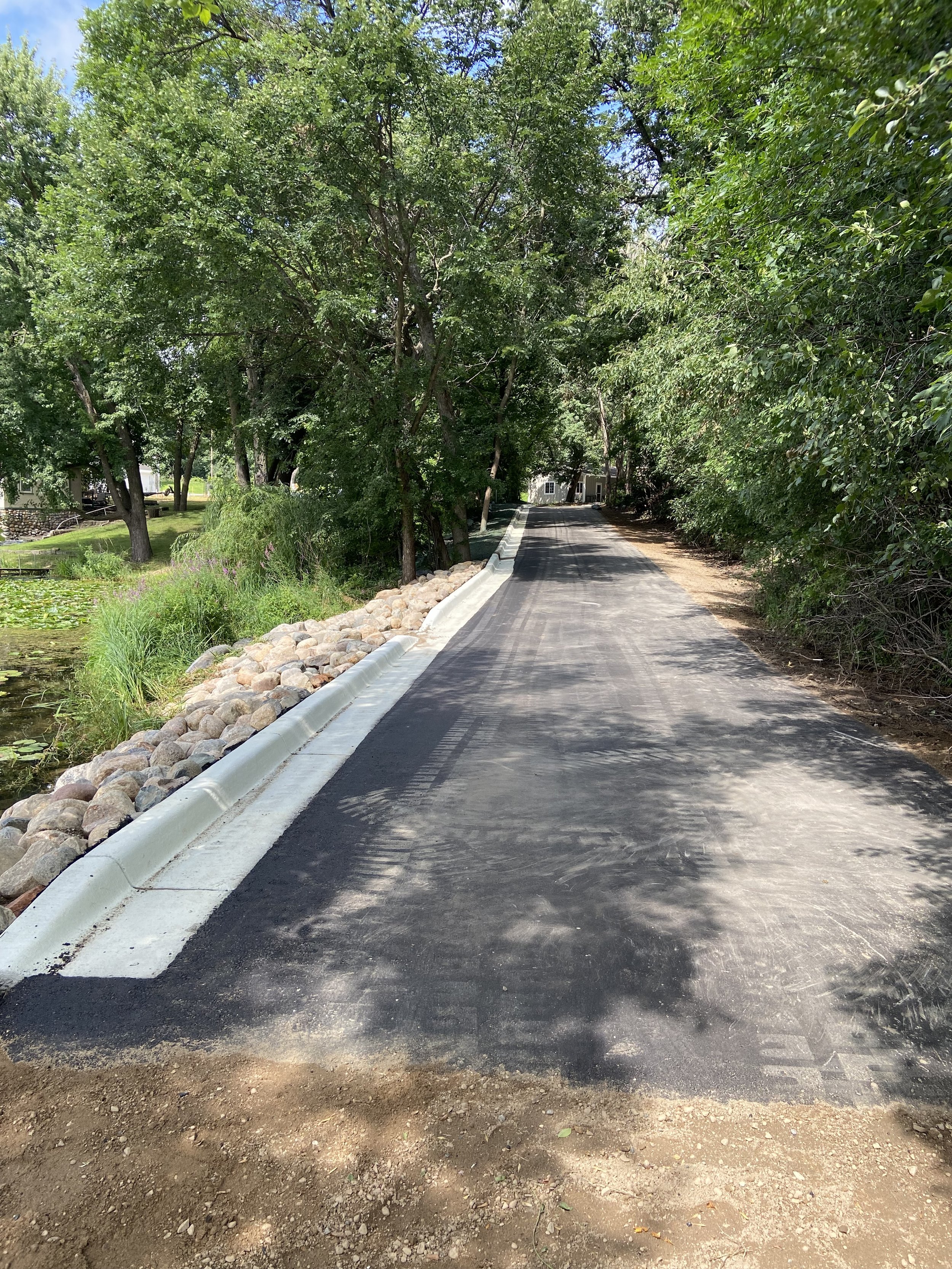 A freshly paved asphalt road bordered by rocks and trees, with a house visible in the distance amid greenery.
