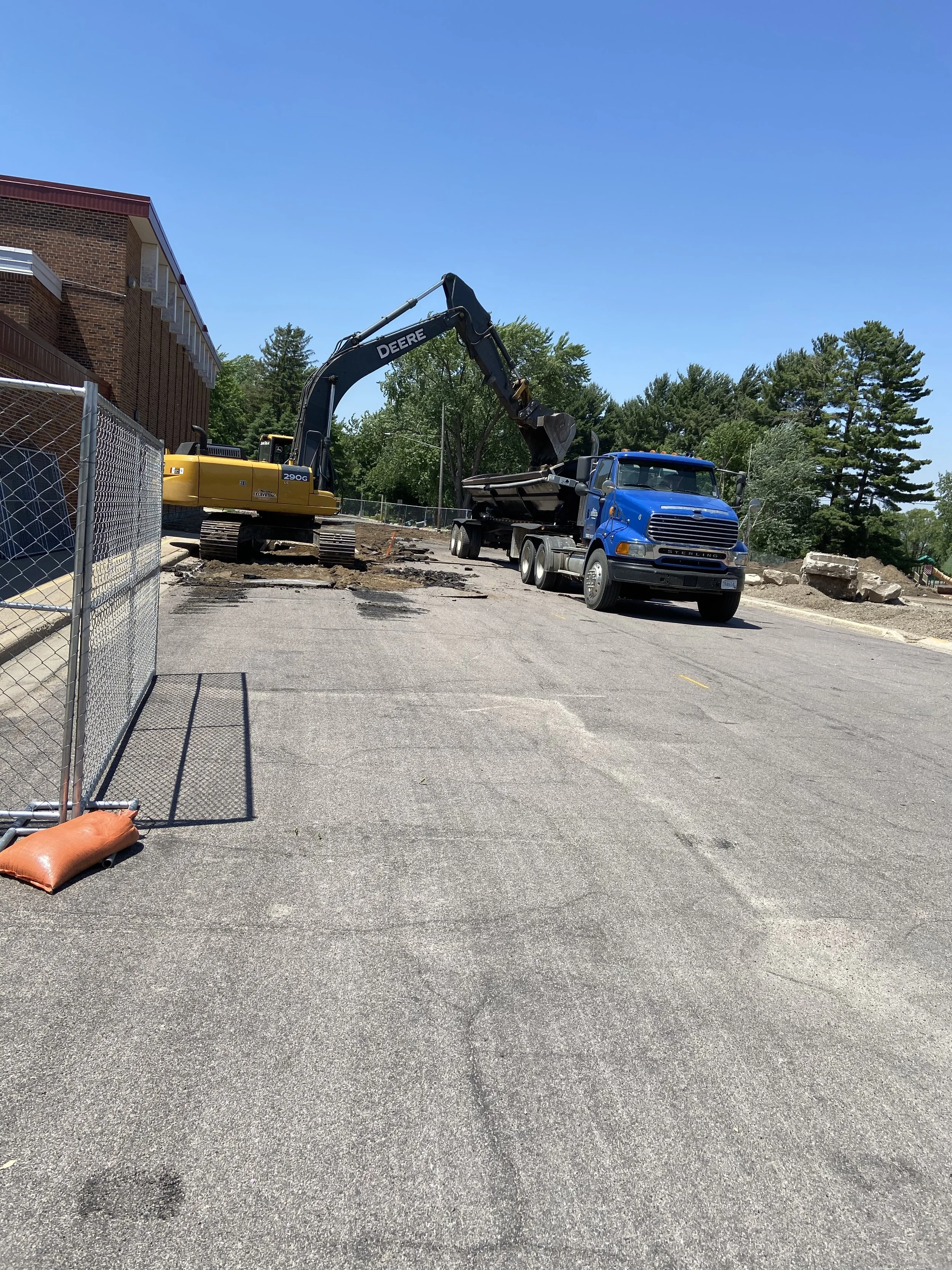 Construction scene with a yellow excavator loading debris into a blue dump truck on a paved area, with a fence and trees in the background.