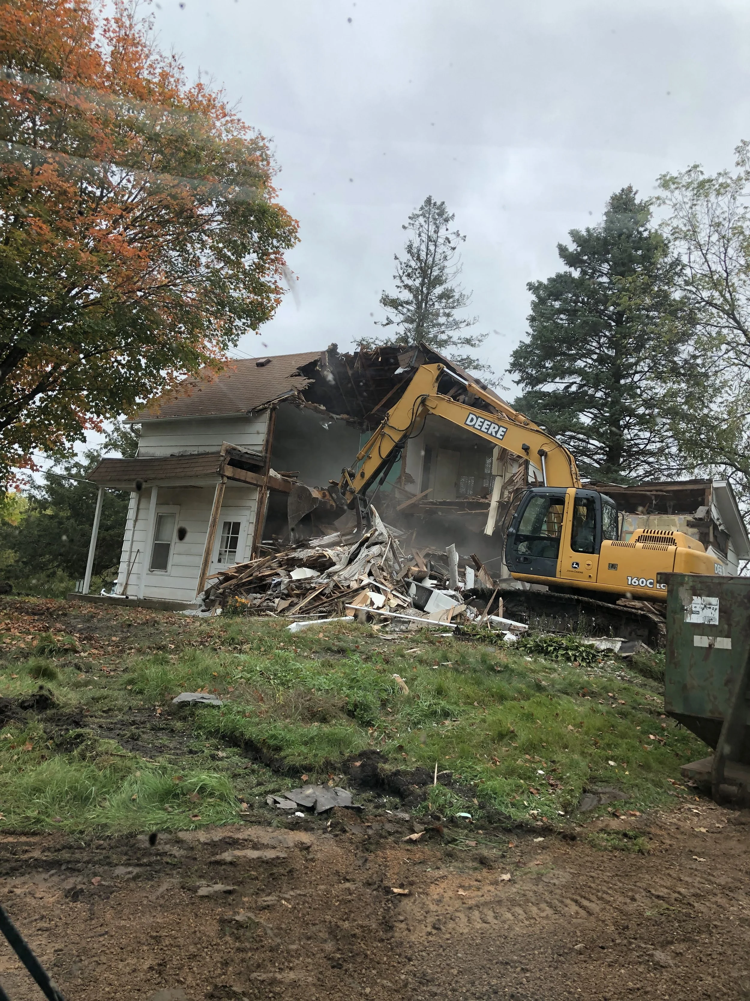 A house being demolished with a large yellow excavator. The house is partially torn down, with debris and wood scattered. Surrounding trees indicate an autumn setting.