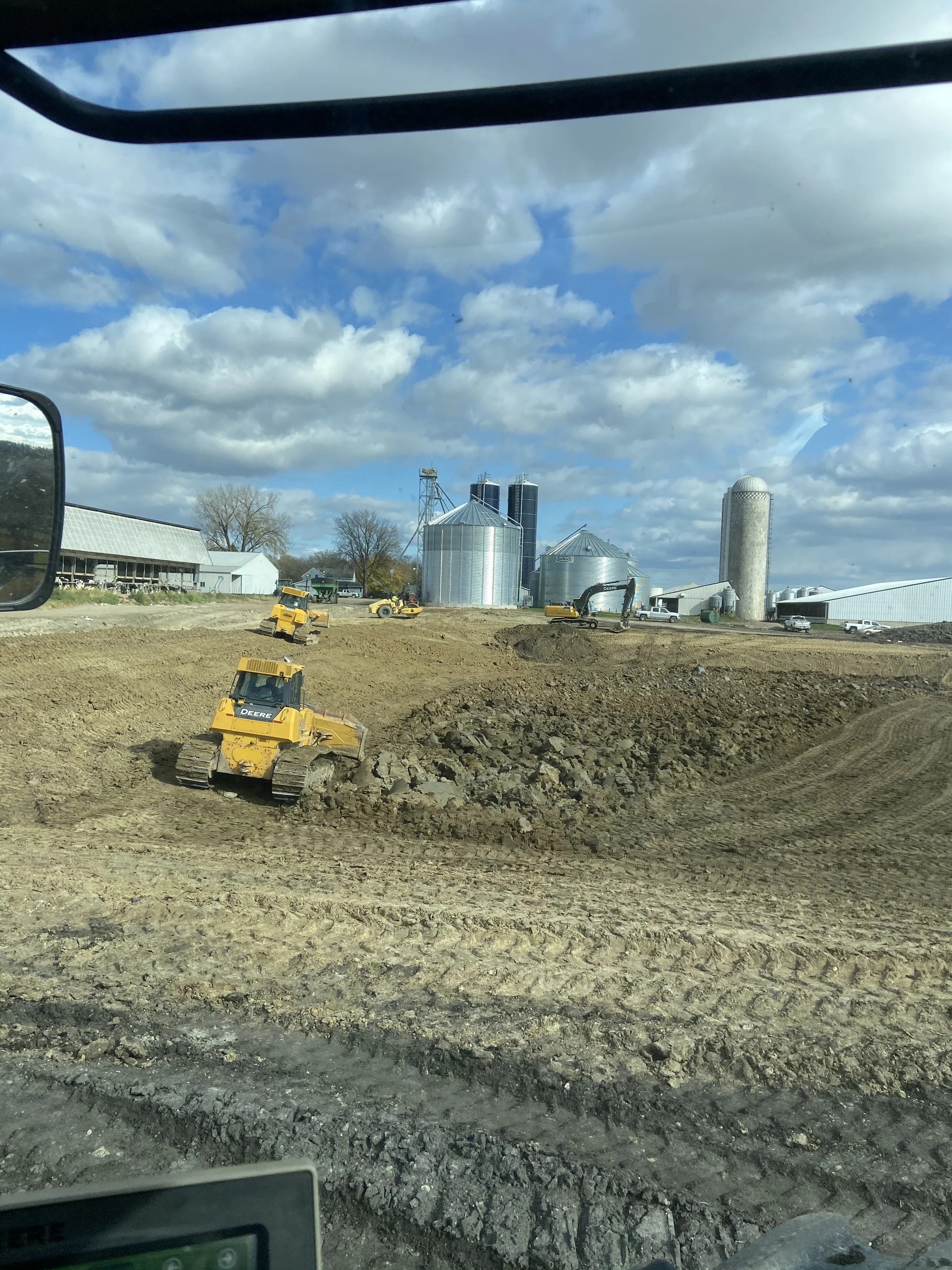 View from inside a construction vehicle showing multiple yellow bulldozers working on a dirt field, with a farm with silos and storage buildings in the background and blue sky with clouds overhead.