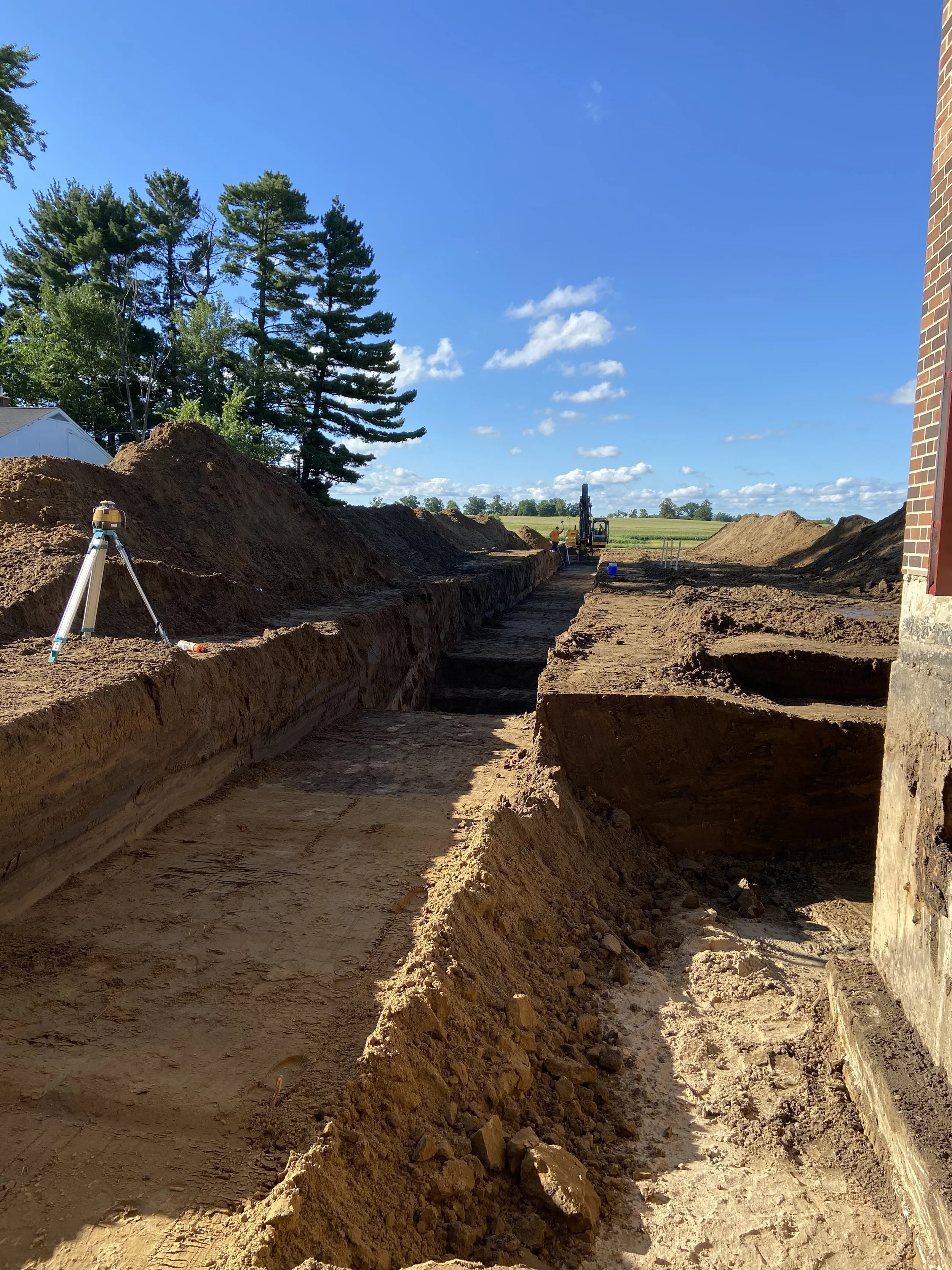 Excavation site with trenches dug in dirt, construction equipment in the background, and a blue sky with scattered clouds.