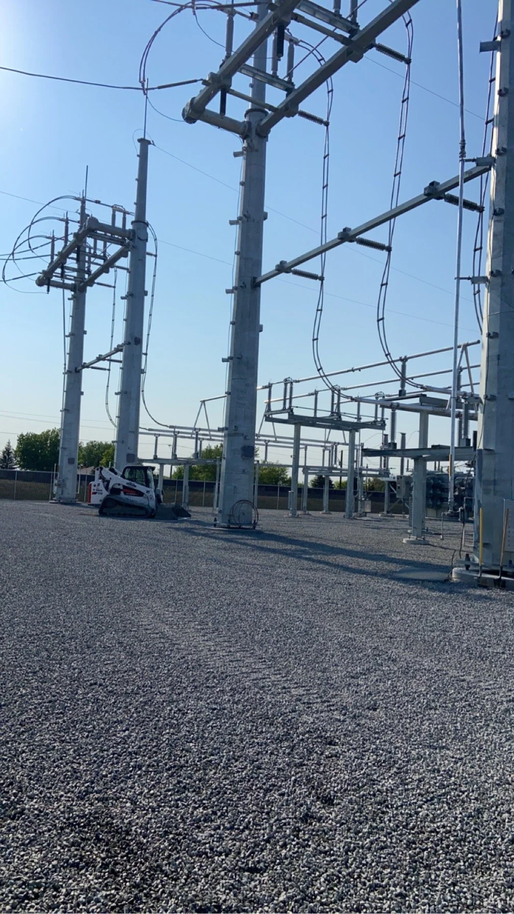 An electrical substation with tall power transmission towers and a small tracked vehicle parked on gravel ground.