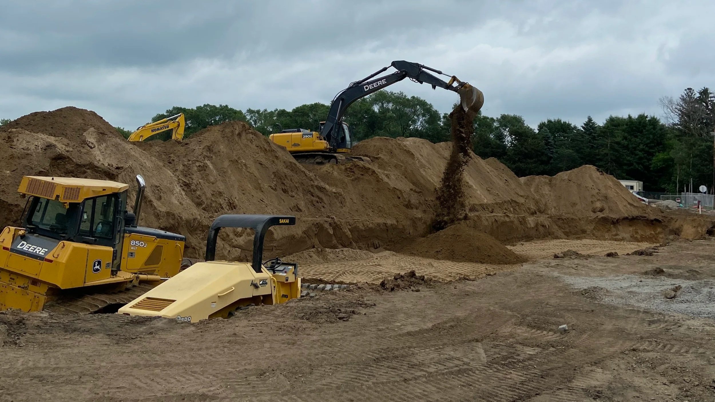 Construction site with large excavator pouring dirt onto a pile, other construction equipment visible, and trees in the background.