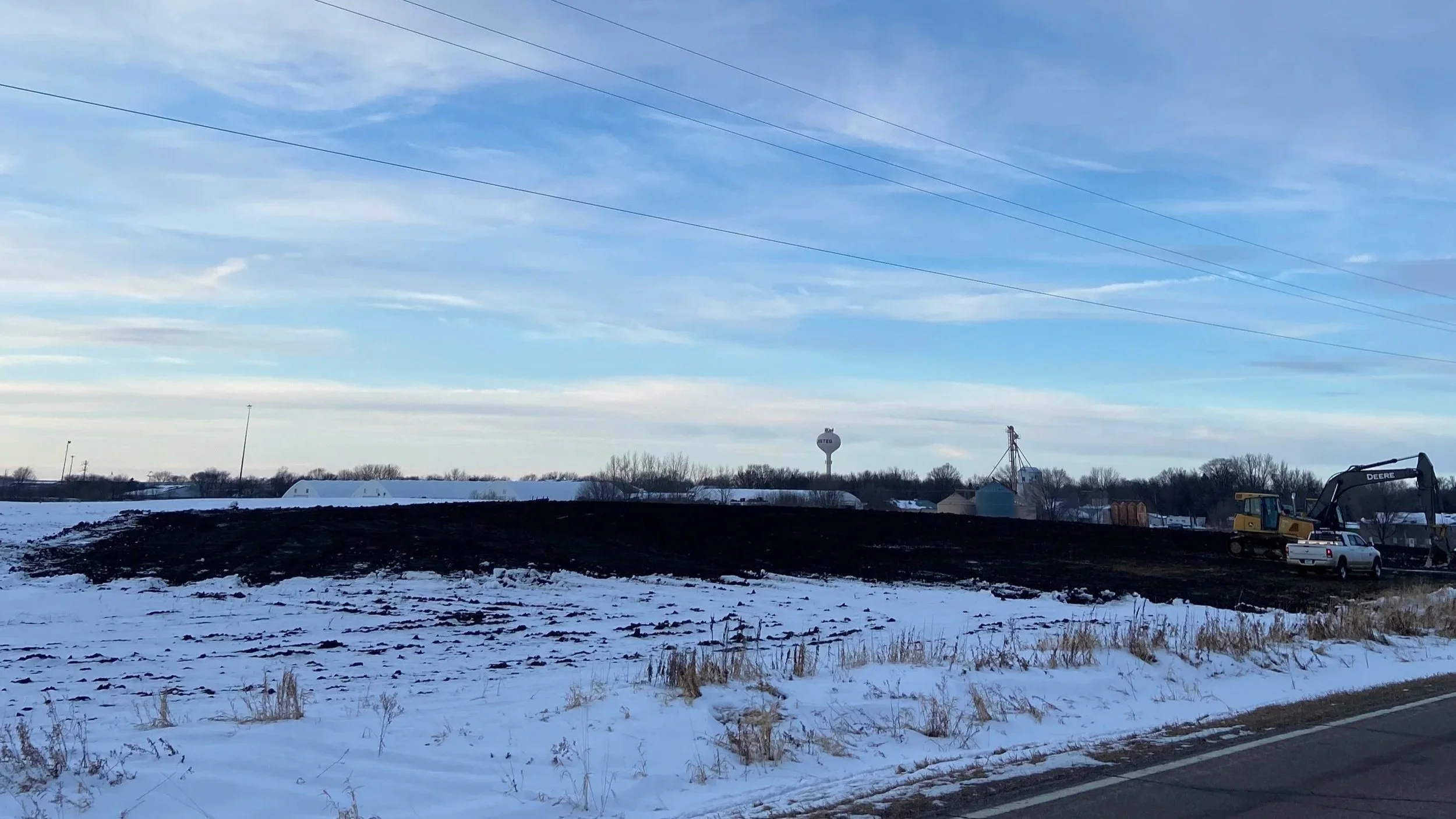 Snow-covered field with a patch of freshly turned dark soil, construction equipment, a white pickup truck, and a water tower in the distance on a partly cloudy winter day.
