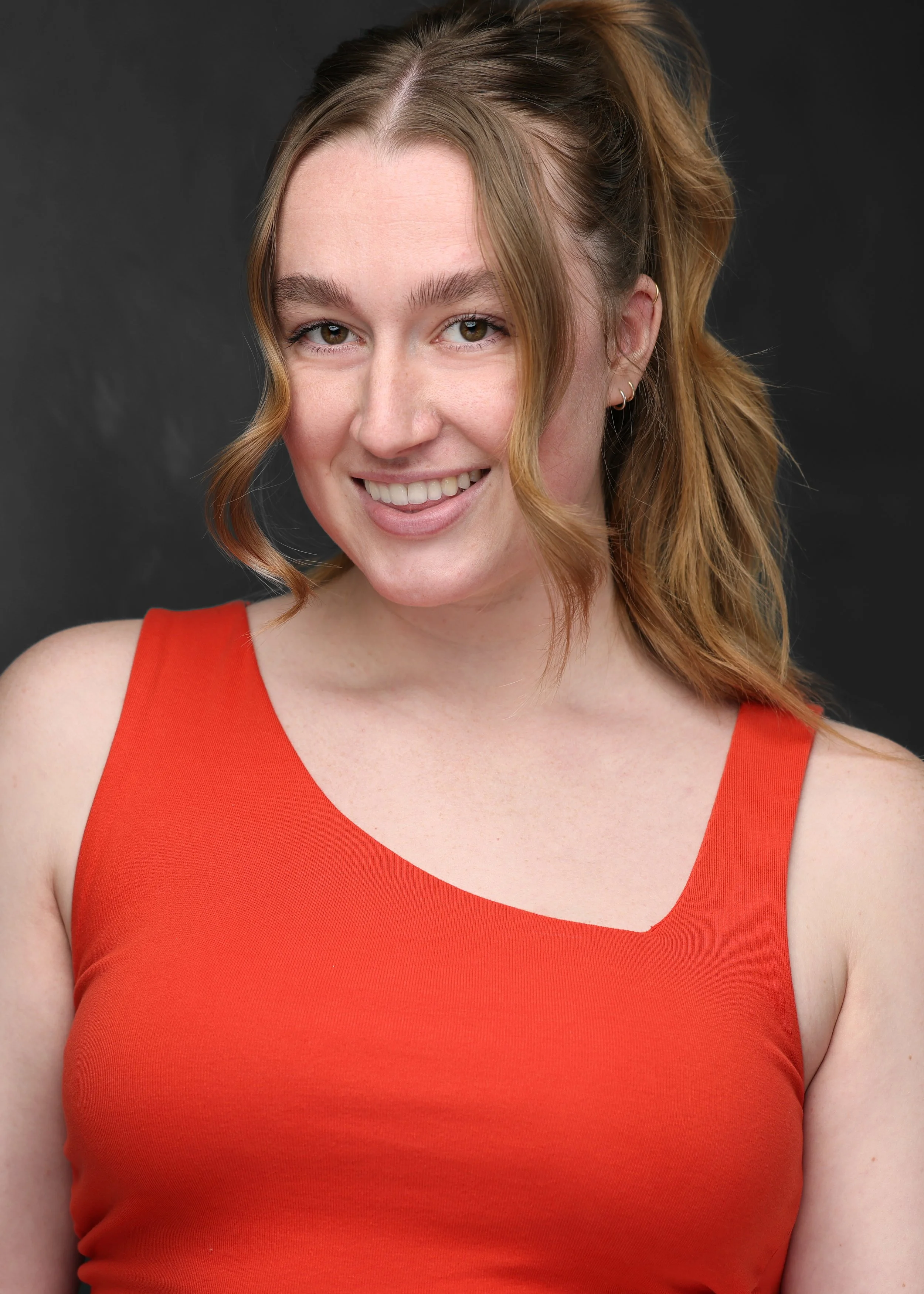 A woman with red hair styled in loose waves, wearing a sleeveless orange top, smiling against a dark background.