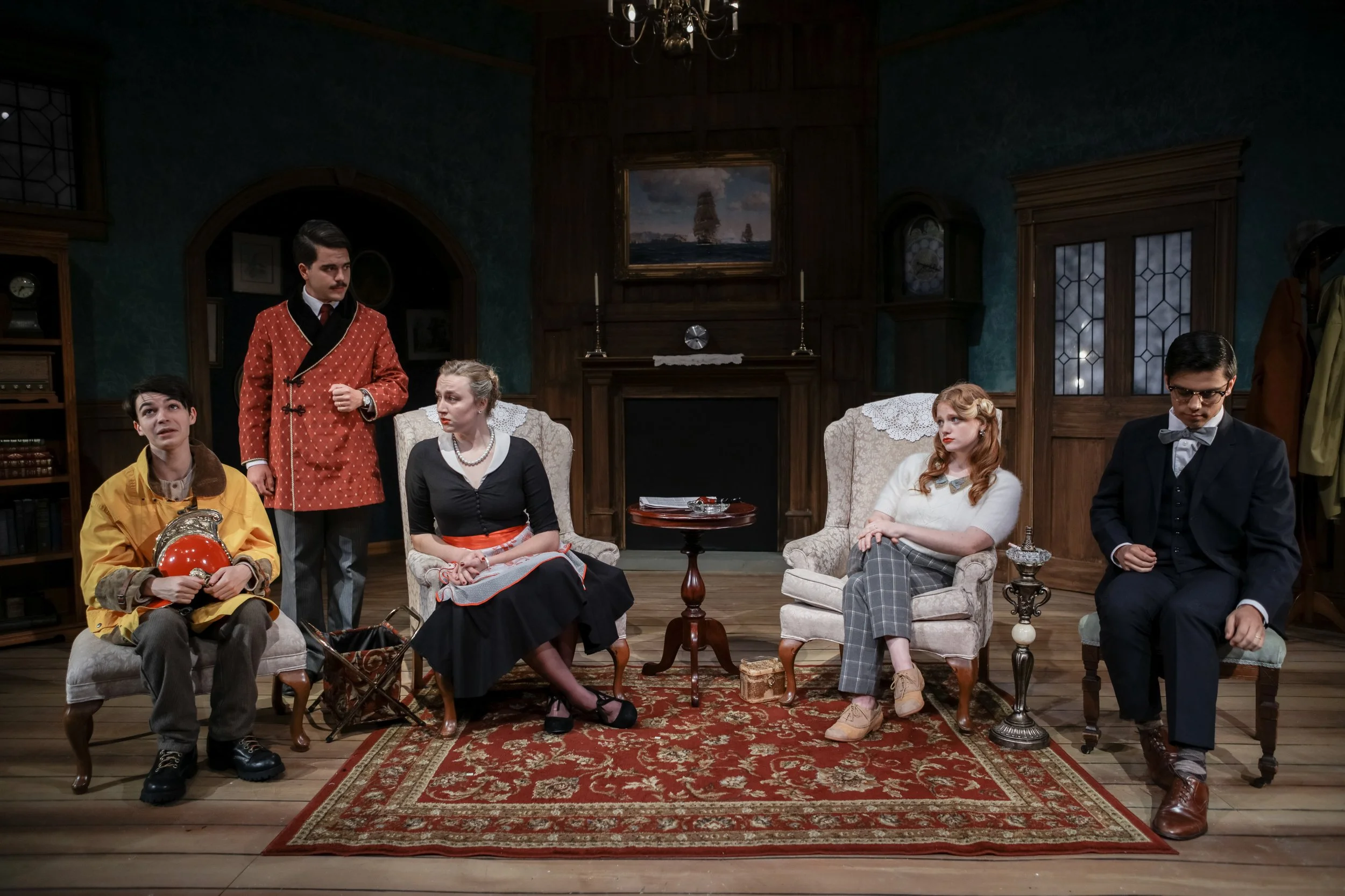 Scene from a theatrical play set in a vintage living room with six actors in period costumes, sitting and standing around a patterned rug, with wooden furniture and a fireplace in the background.