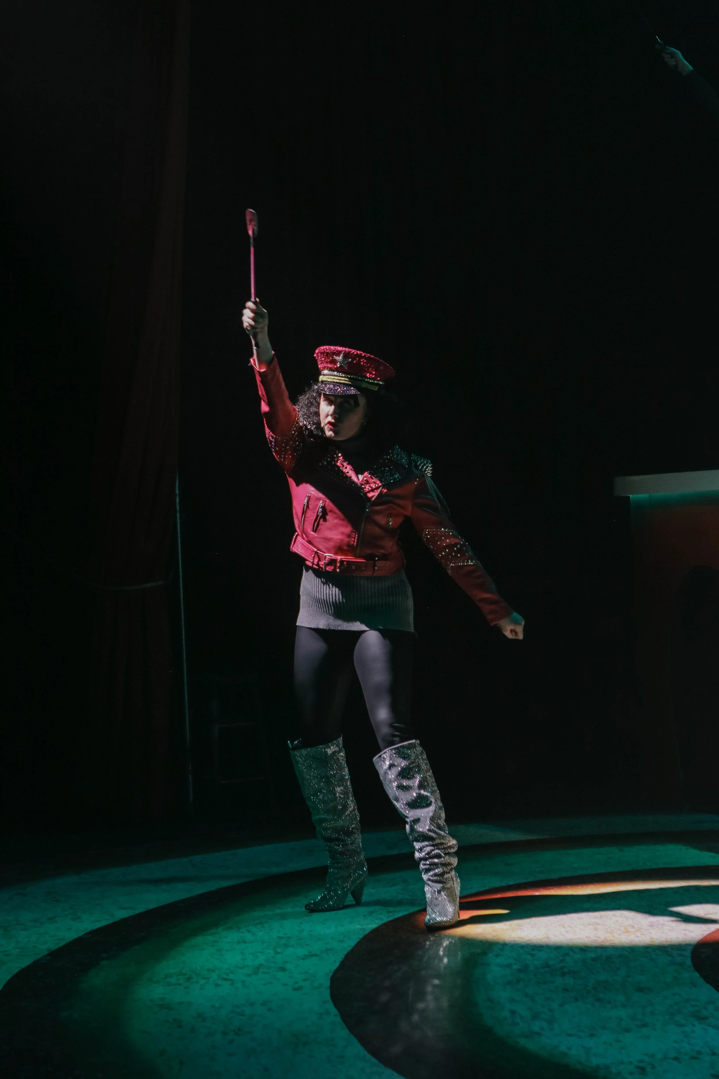 Performer in a glittery military-style hat, bright jacket, and shiny boots holding a baton on a spotlighted stage.