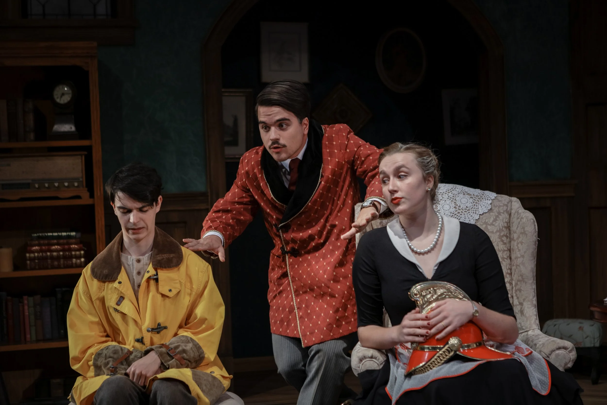 Three actors on stage in a play, with one standing and gesturing, and two seated, in vintage costumes in a room with wooden furniture and framed pictures.