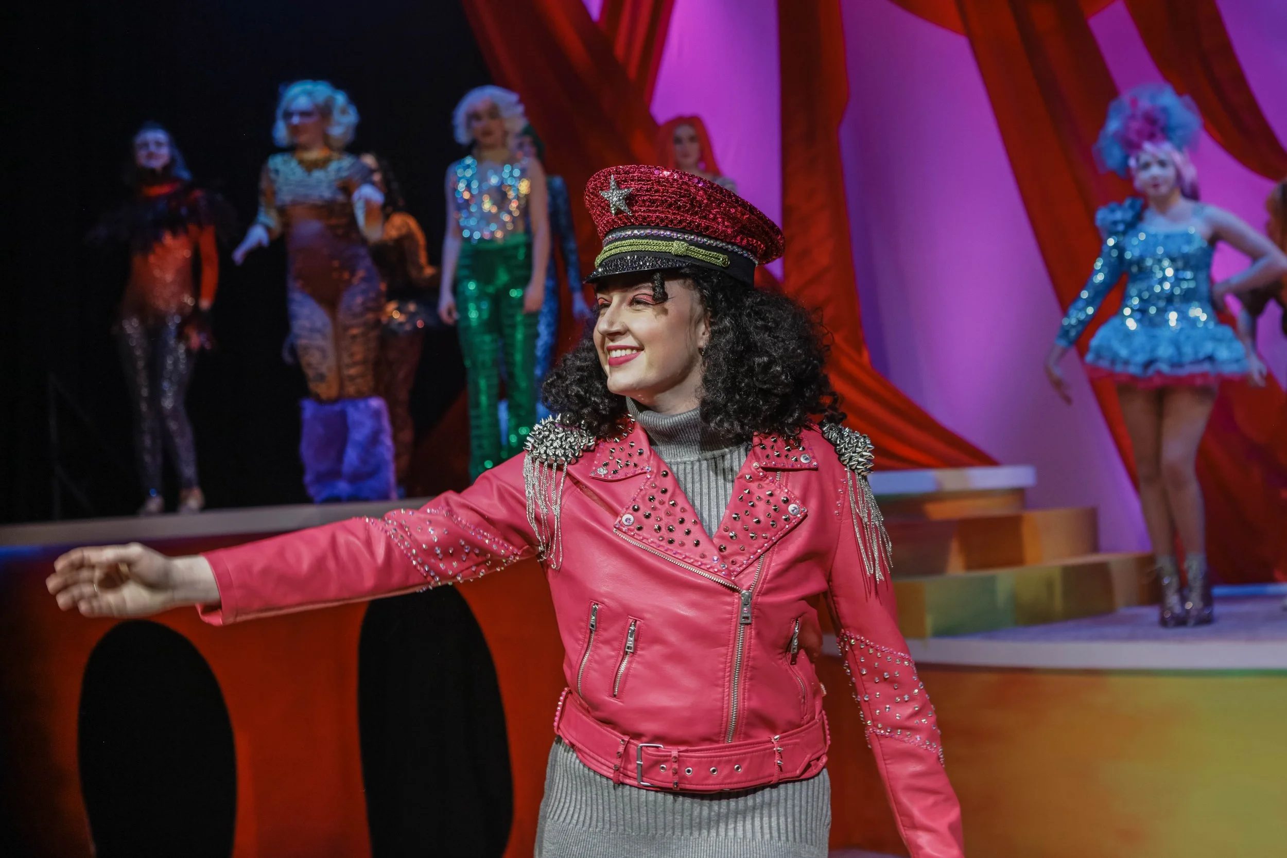 A woman with curly black hair wearing a pink leather jacket with silver studs, a gray turtleneck, and a glittery hat with a star, standing in front of a colorful stage with performers in vibrant costumes.